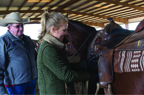 A 4-her prepares her saddle on a quarter horse while instructor watches.