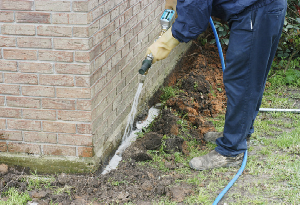 A person in protective clothing and gloves pours liquid termiticide into a trench around the base of a brick building.