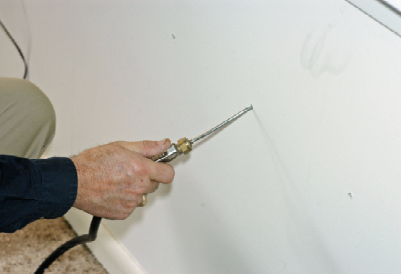 A person's hand with a long, narrow tool inserted into a sheetrock wall.