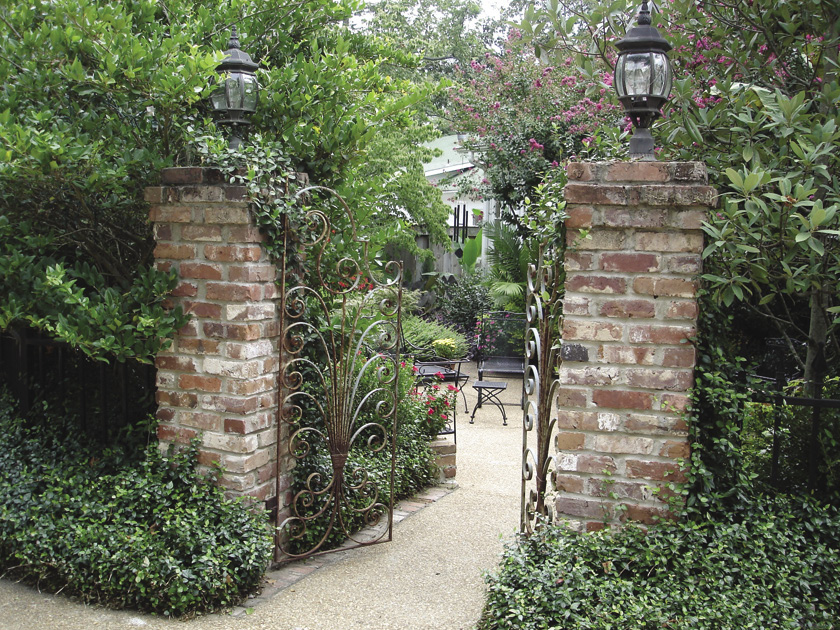 A wrought-iron gate supported by two brick pillars with black light fixtures on top. The gate is surrounded by green foliage and shows a view of a brick patio in a garden.