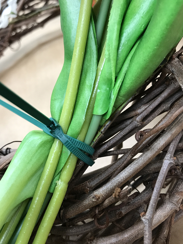 Green stems of fake flowers zip-tied to grapevine wreath.