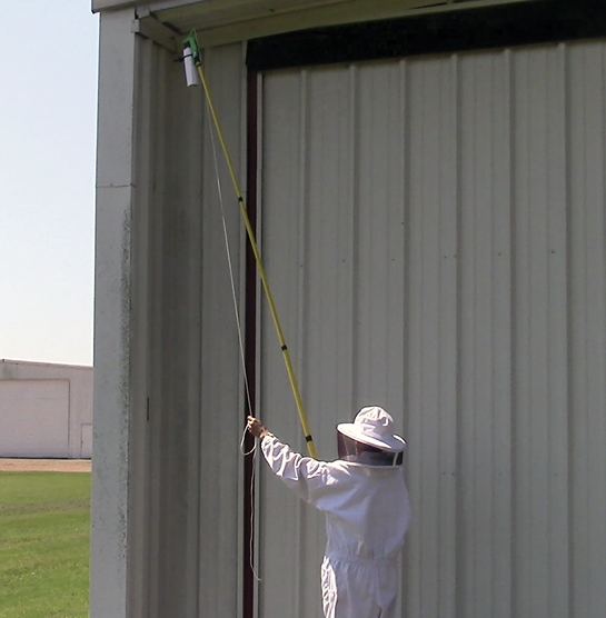 A man holds the long pole and operates the sprayer by pulling a twine to apply pesticide at the top of a tall building.  