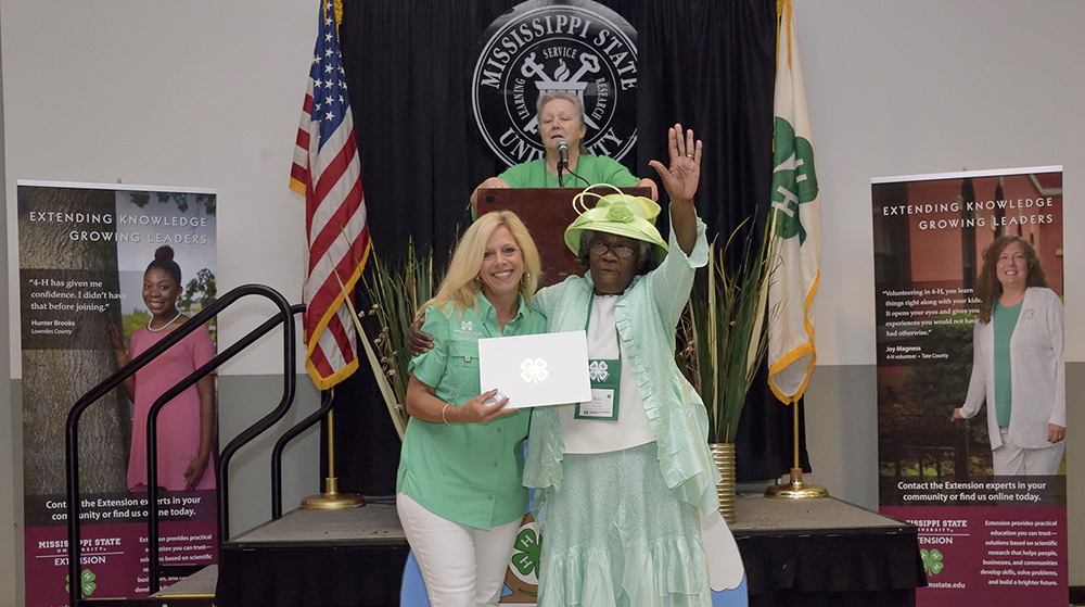Two women, both wearing green, stand smiling in front of a podium featuring a speaker looking on.  The woman on the left holds a white folder with a silver 4-H clover emblazoned in the center.