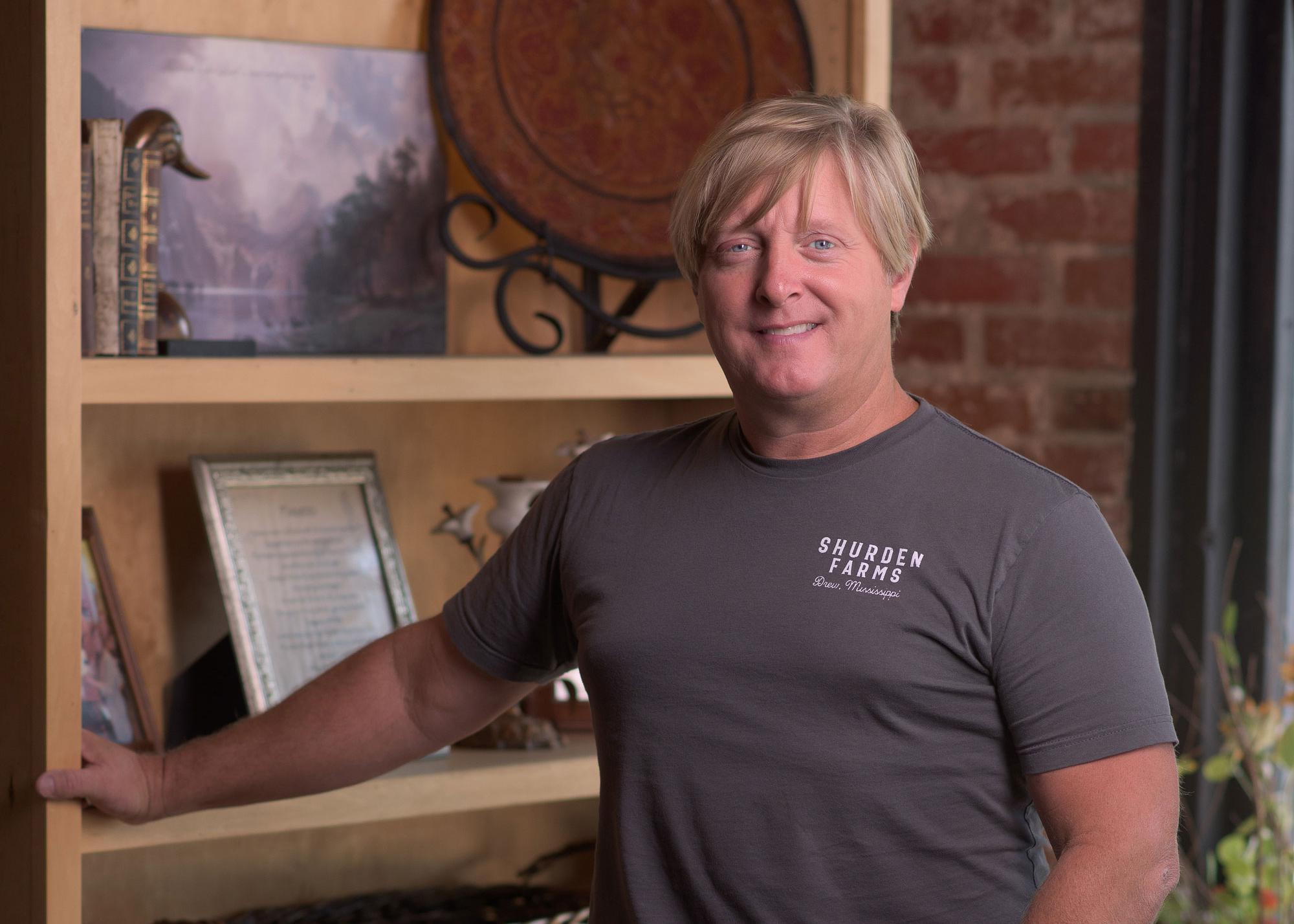 A blonde man wearing a T-shirt with Shuden Farms listed on it standing in front of a bookshelf, smiling.
