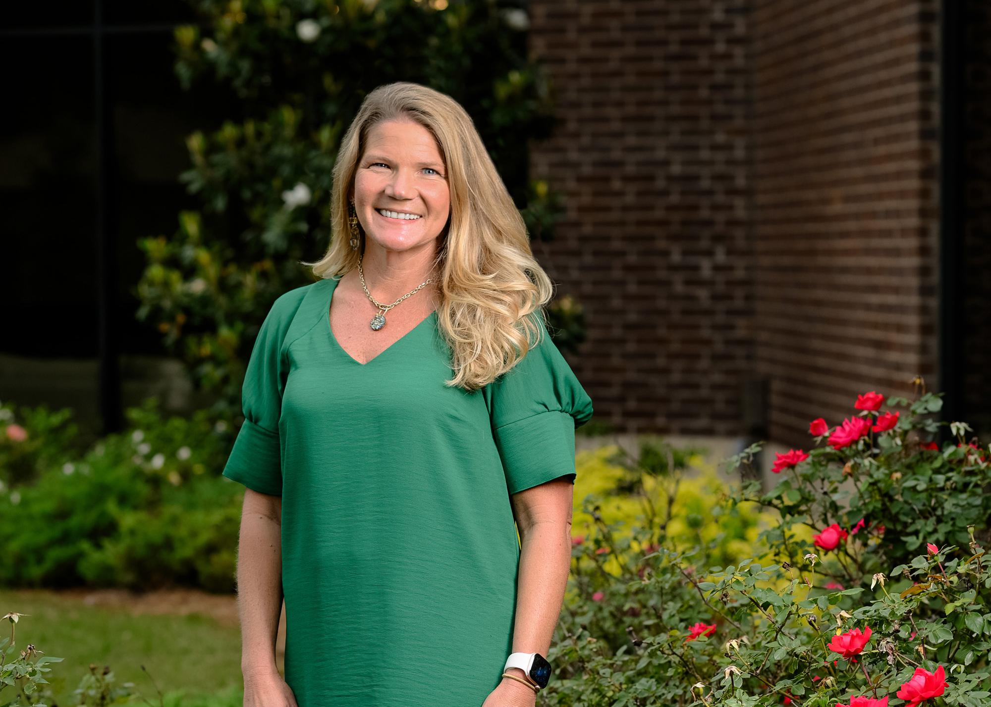 A smiling person wearing green and standing outside in front of roses.