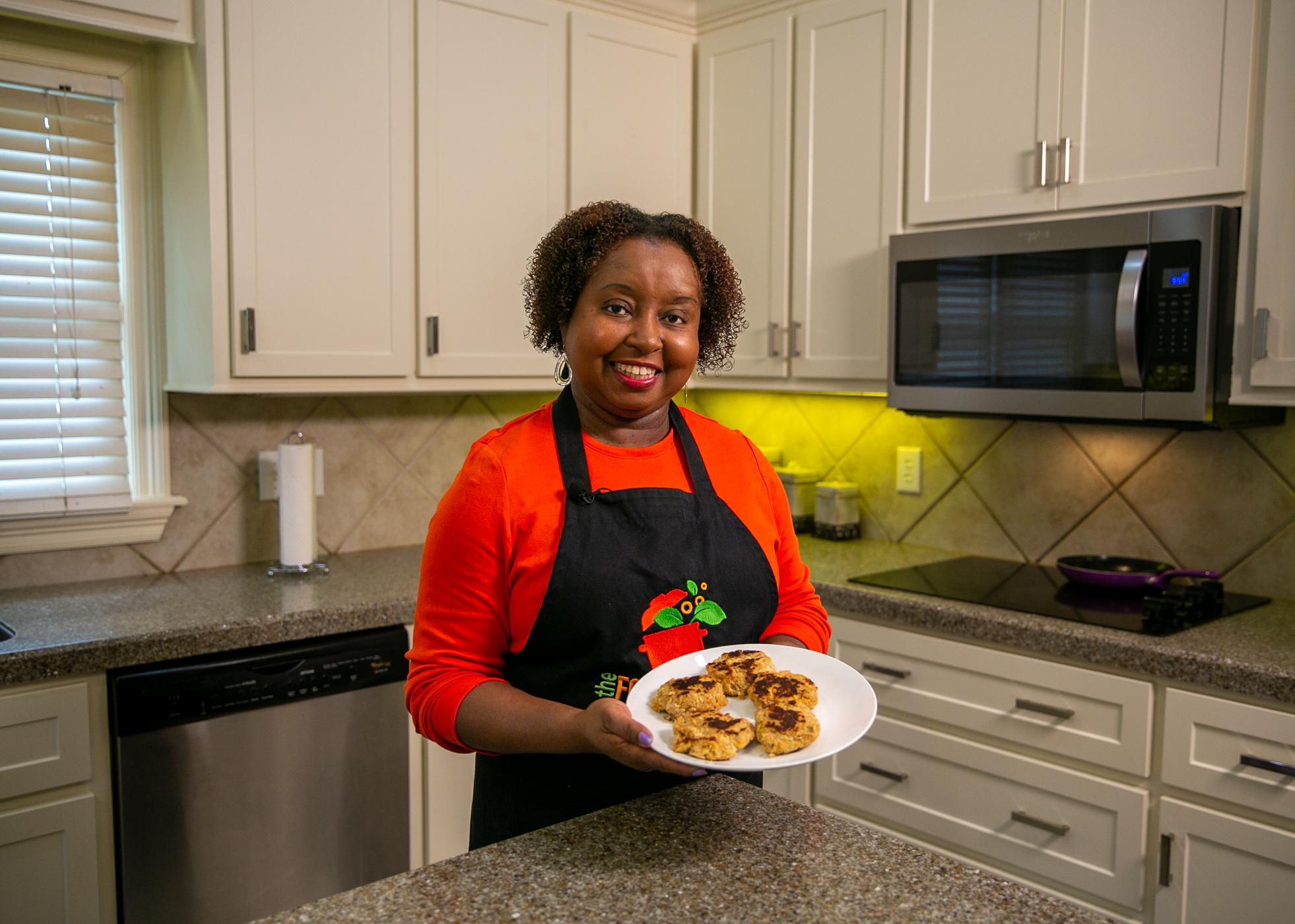 A woman stands in a kitchen holding a plate of salmon patties for the camera.