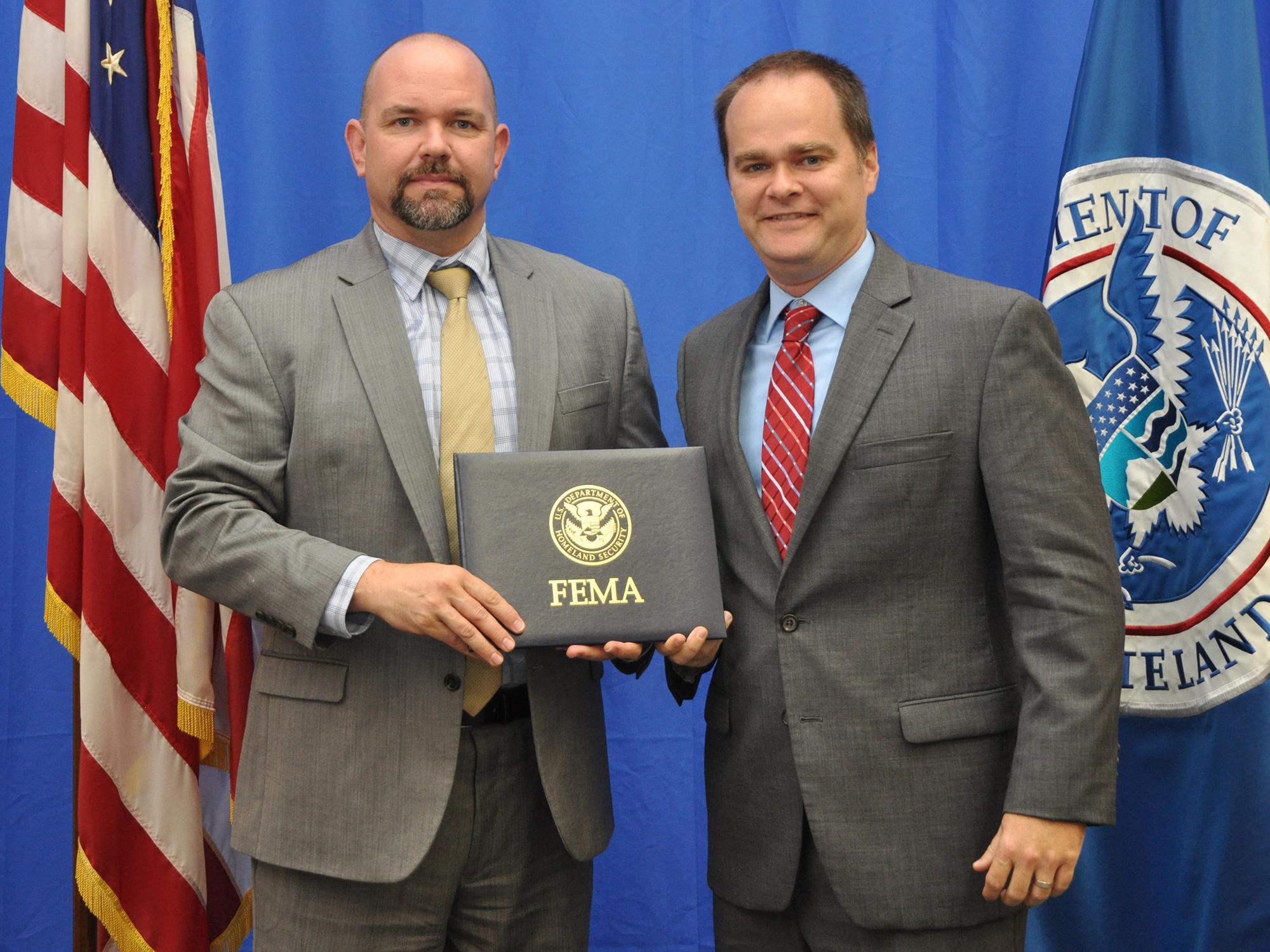 Two men on a stage holding a FEMA certificate and looking at the camera.