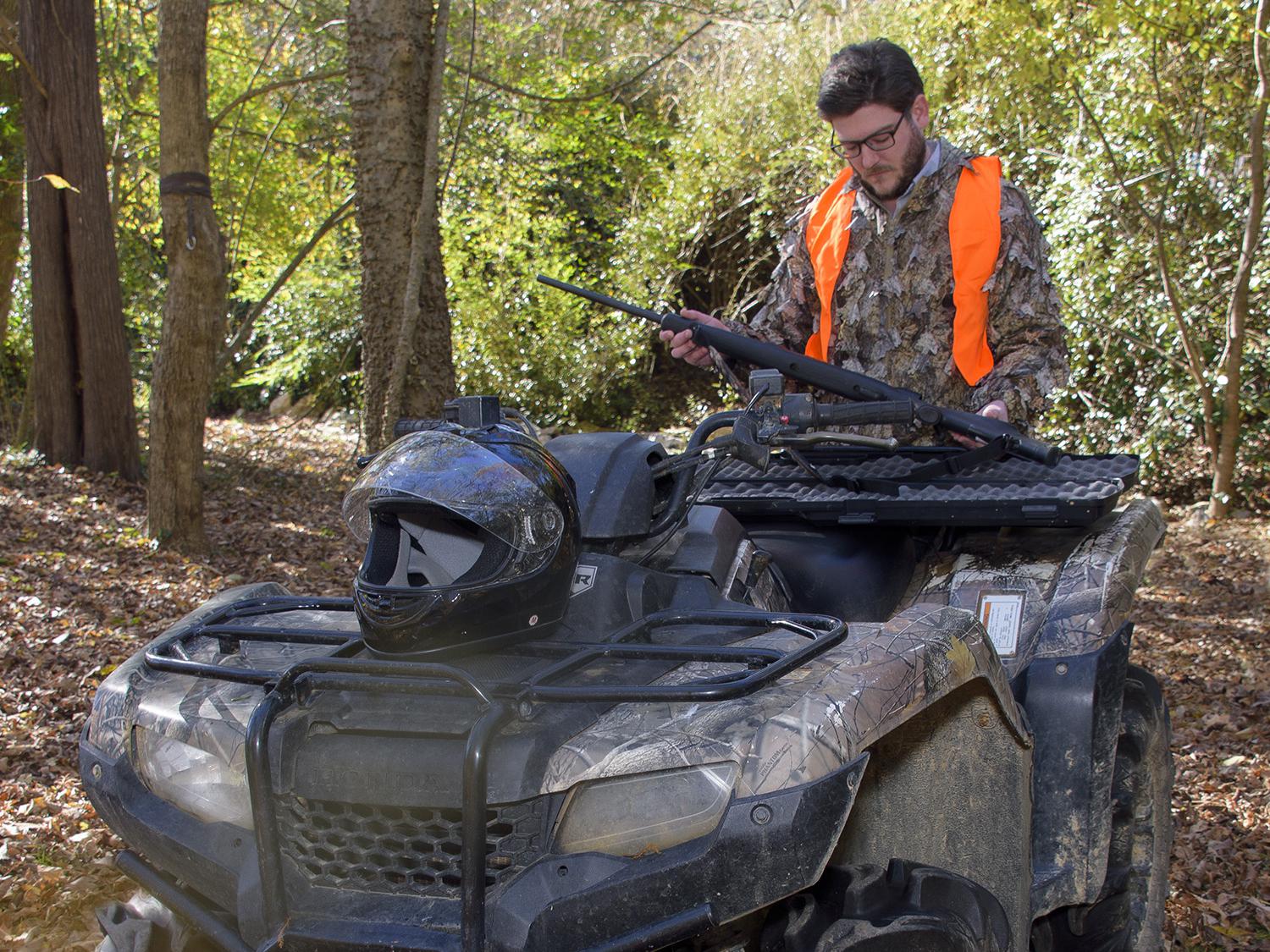 A hunter in camouflage and an orange vest places his rifle into storage on the back of an ATV in the woods.