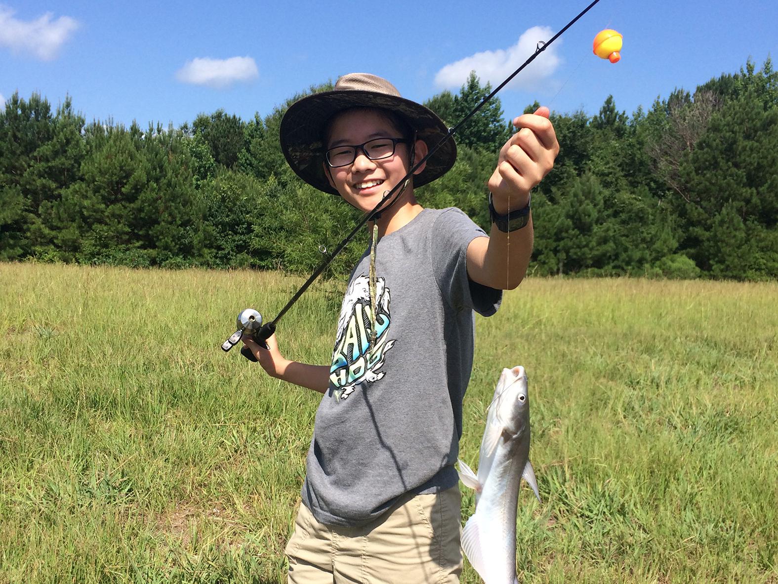 A teenage boy proudly holds up a catfish on his fishing line.