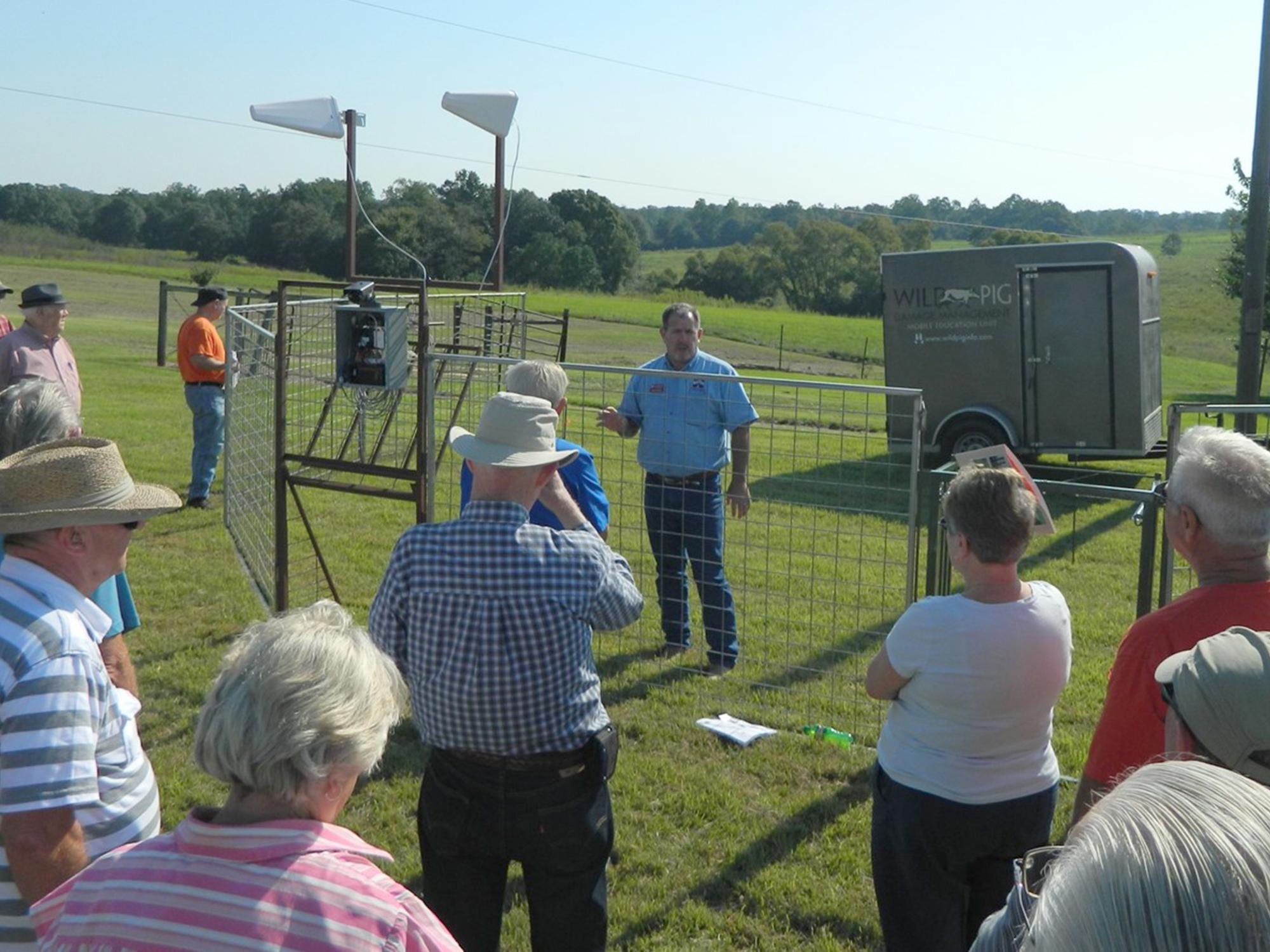 A group of adults gather outside a metal corral with electronics attached to it with a speaker inside the trap.