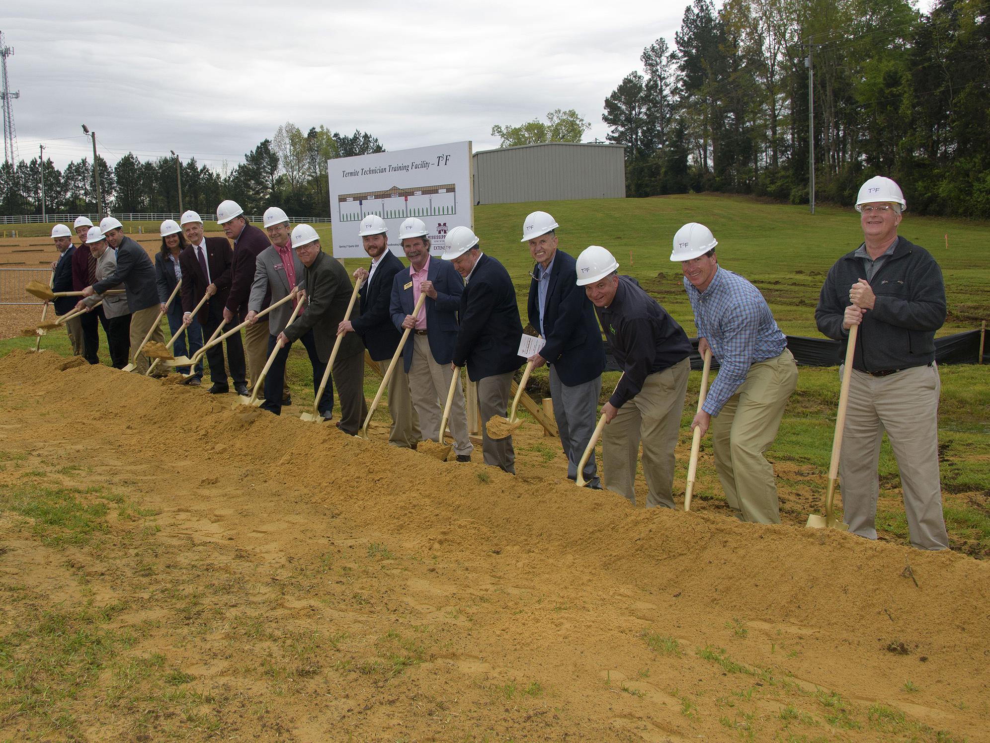 A group of more than a dozen people in hard hats break ground with shovels.