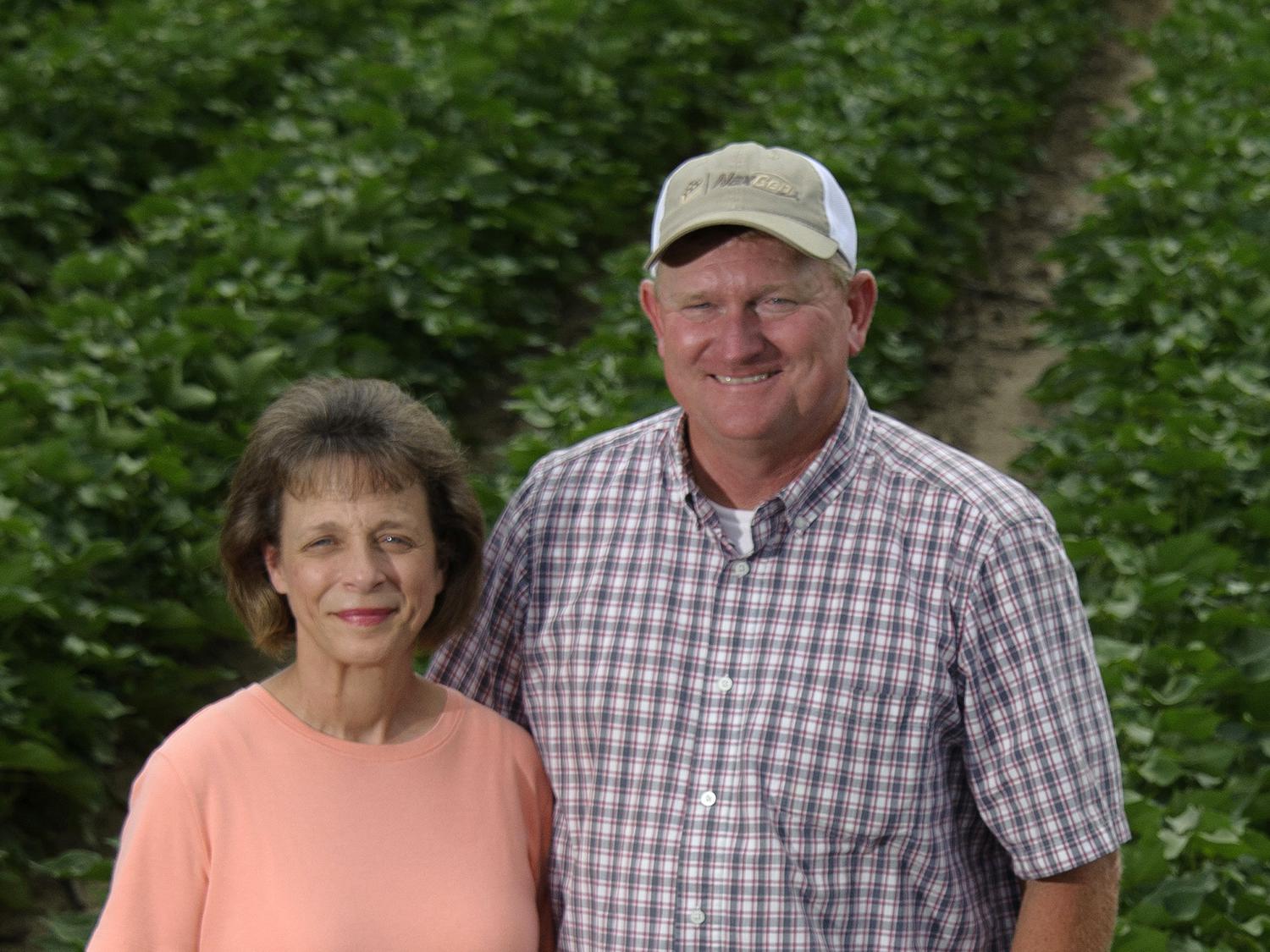 A wife and husband stand in a field with cotton rows.