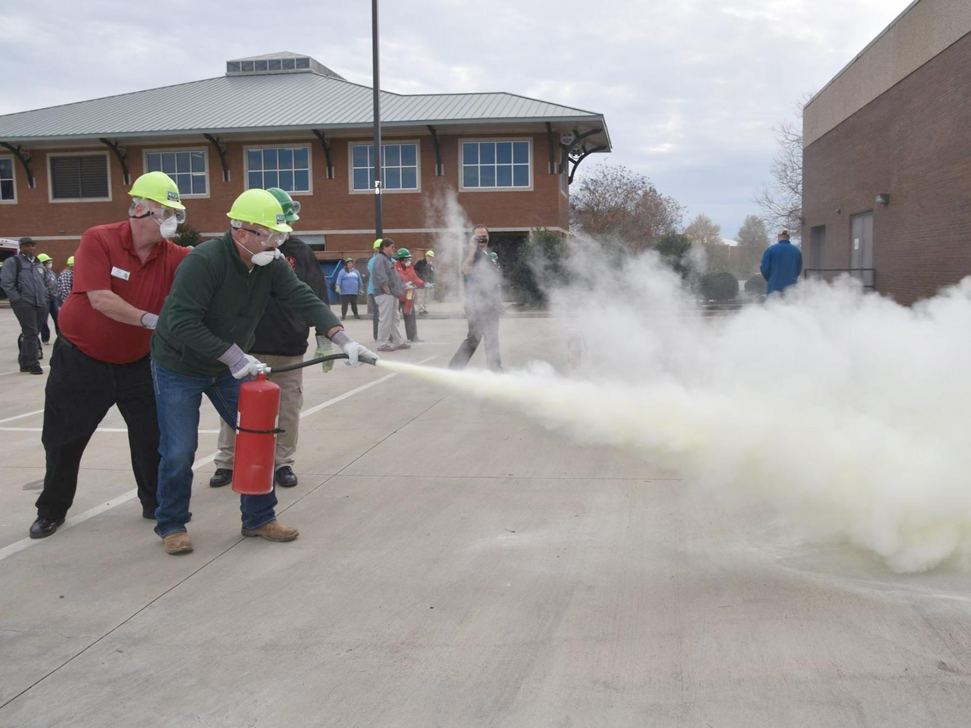 Two men wearing hard hats and masks activate a fire extinguisher as MyPI training participants watch. 