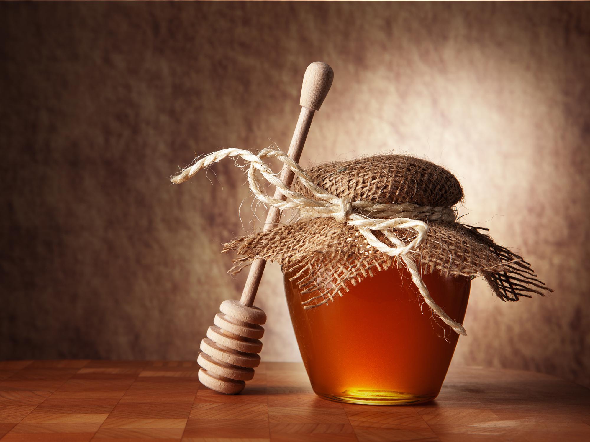 Wooden honey dipper lying against a small jar of honey that is covered with a decorative piece of fabric tied with a string.