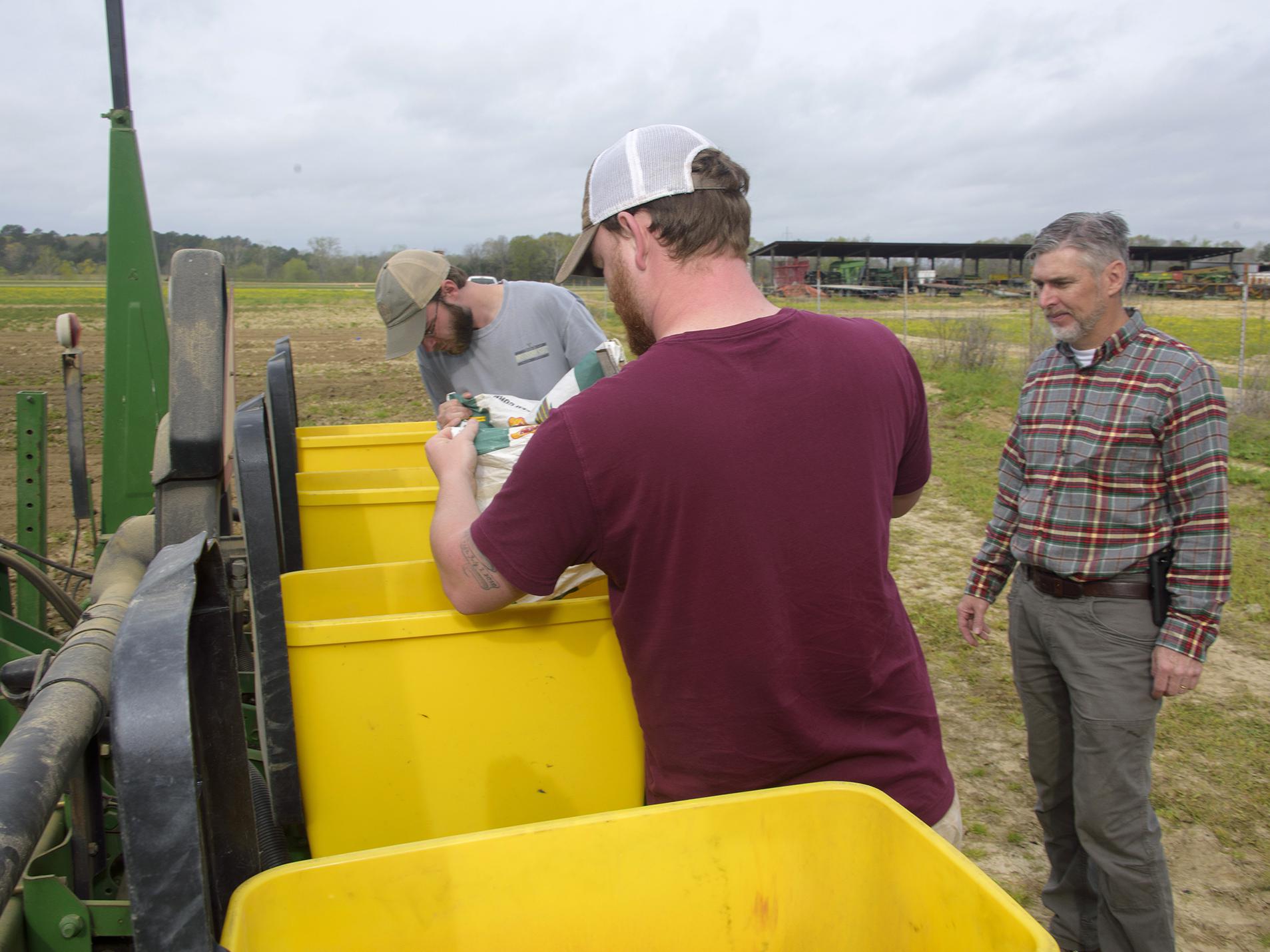 Two young men pour seed into bright yellow bins while a man watches.
