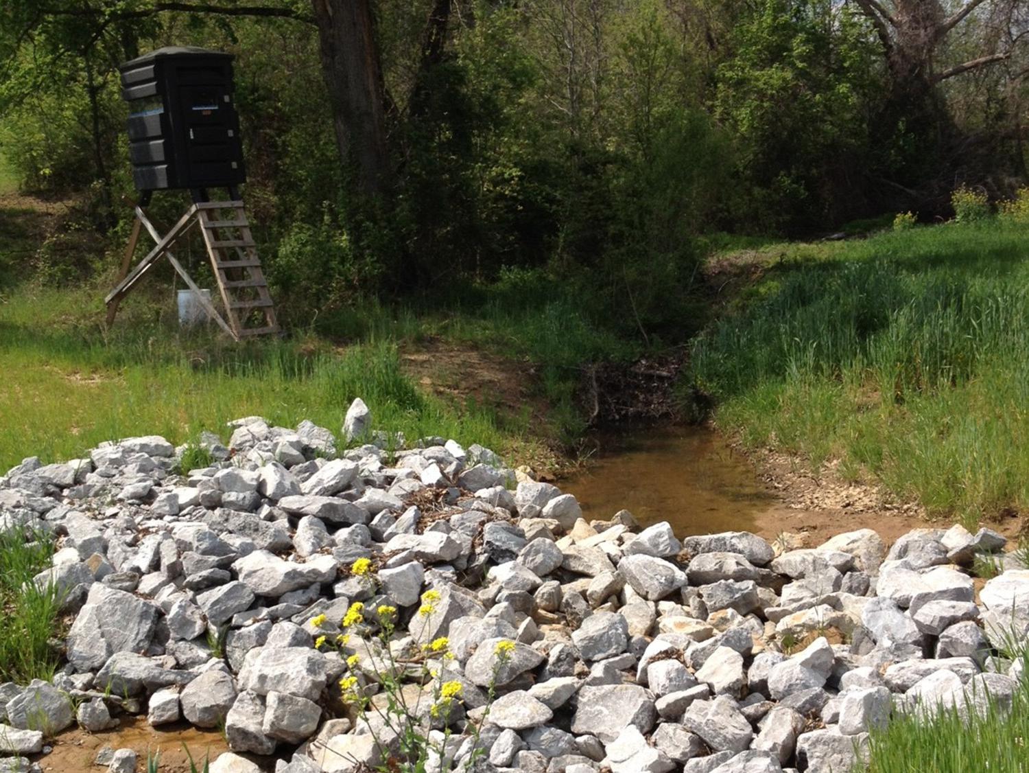 A pile of large gray rocks stretches across a ditch in a country setting.