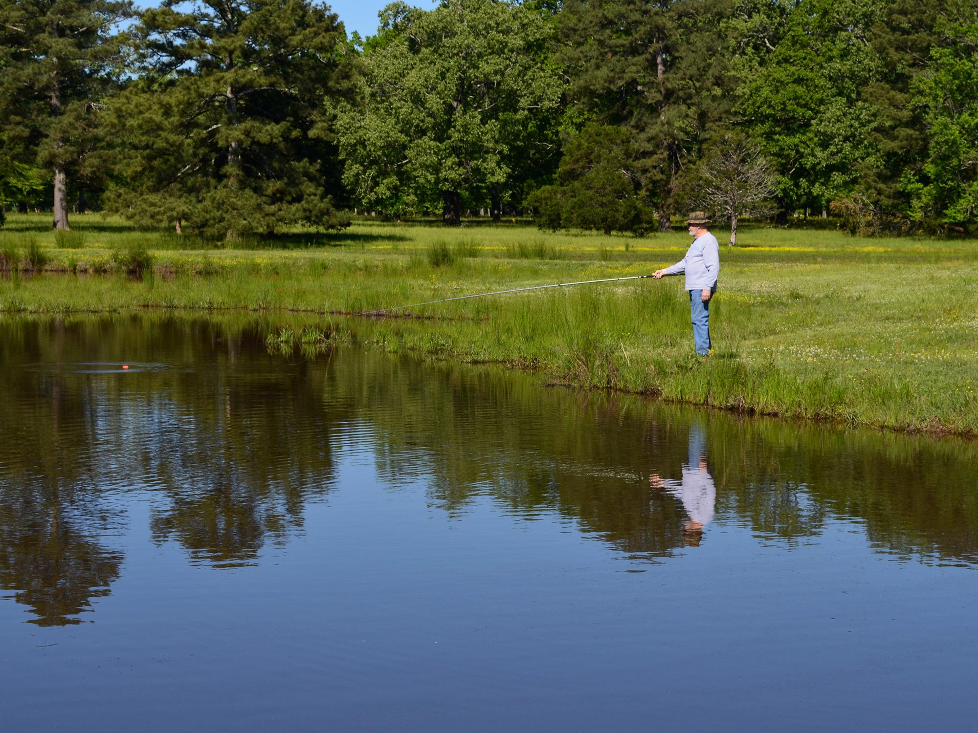 Older man holds a fishing pole on the shoreline while watching a red cork on a quiet pond.