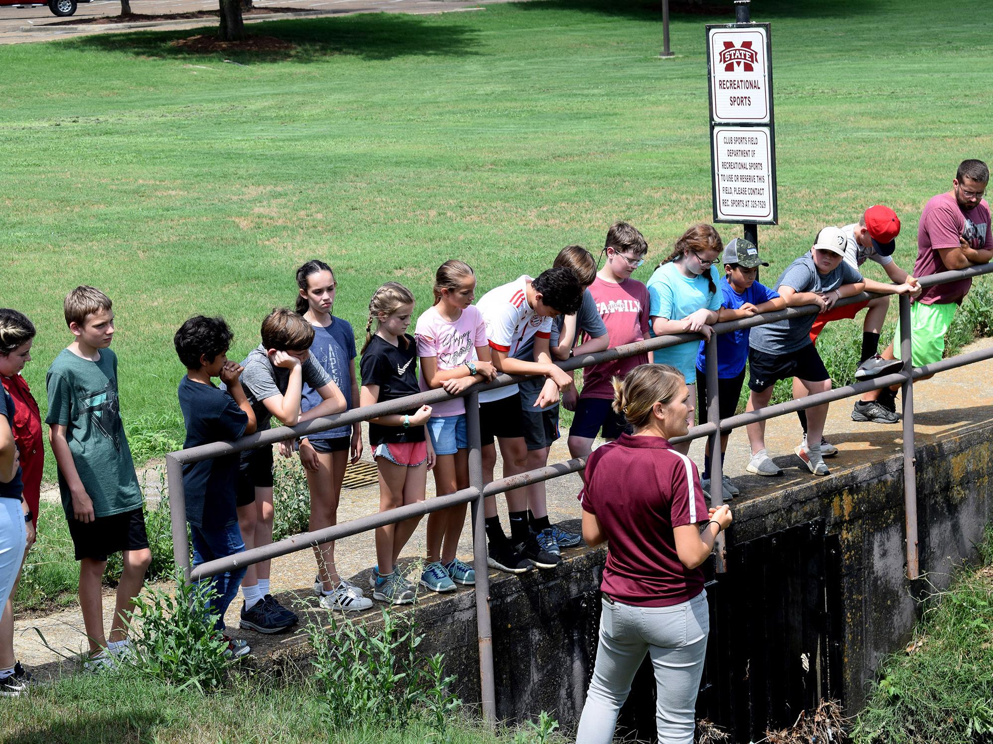 More than a dozen young people listen to a teacher while they stand on a cement bridge overlooking a large ditch.