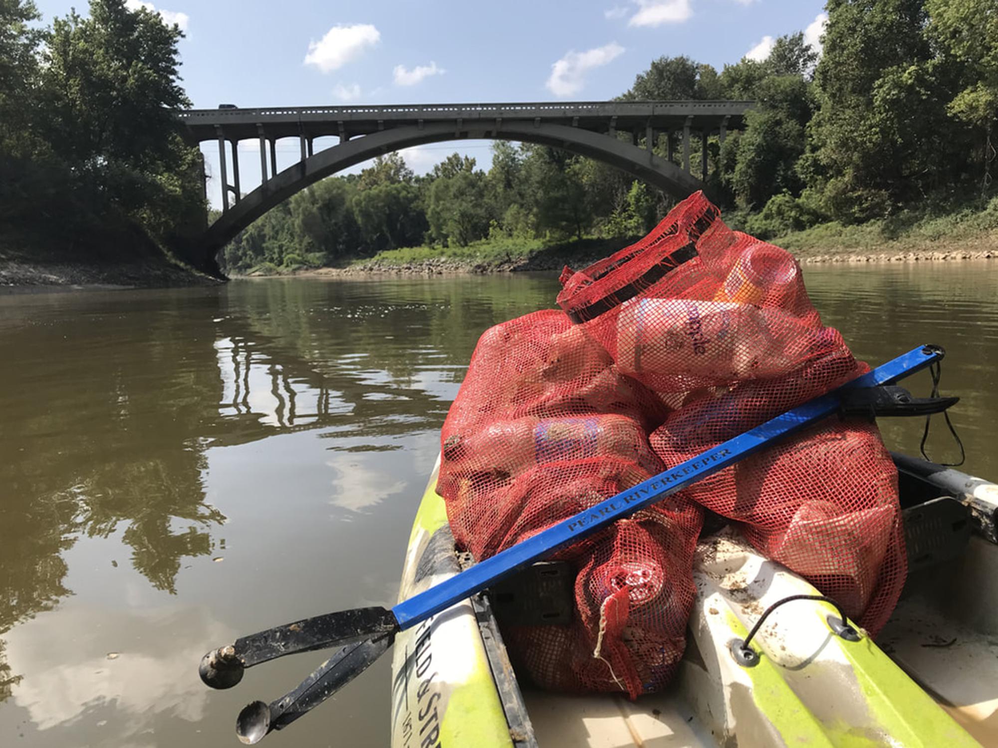 The front of a kayak has two red meshed bags containing litter and a blue trash-grabbing tool with the words “Pearl Riverkeeper” printed on it. The boat is on a small river and approaching a highway bridge.