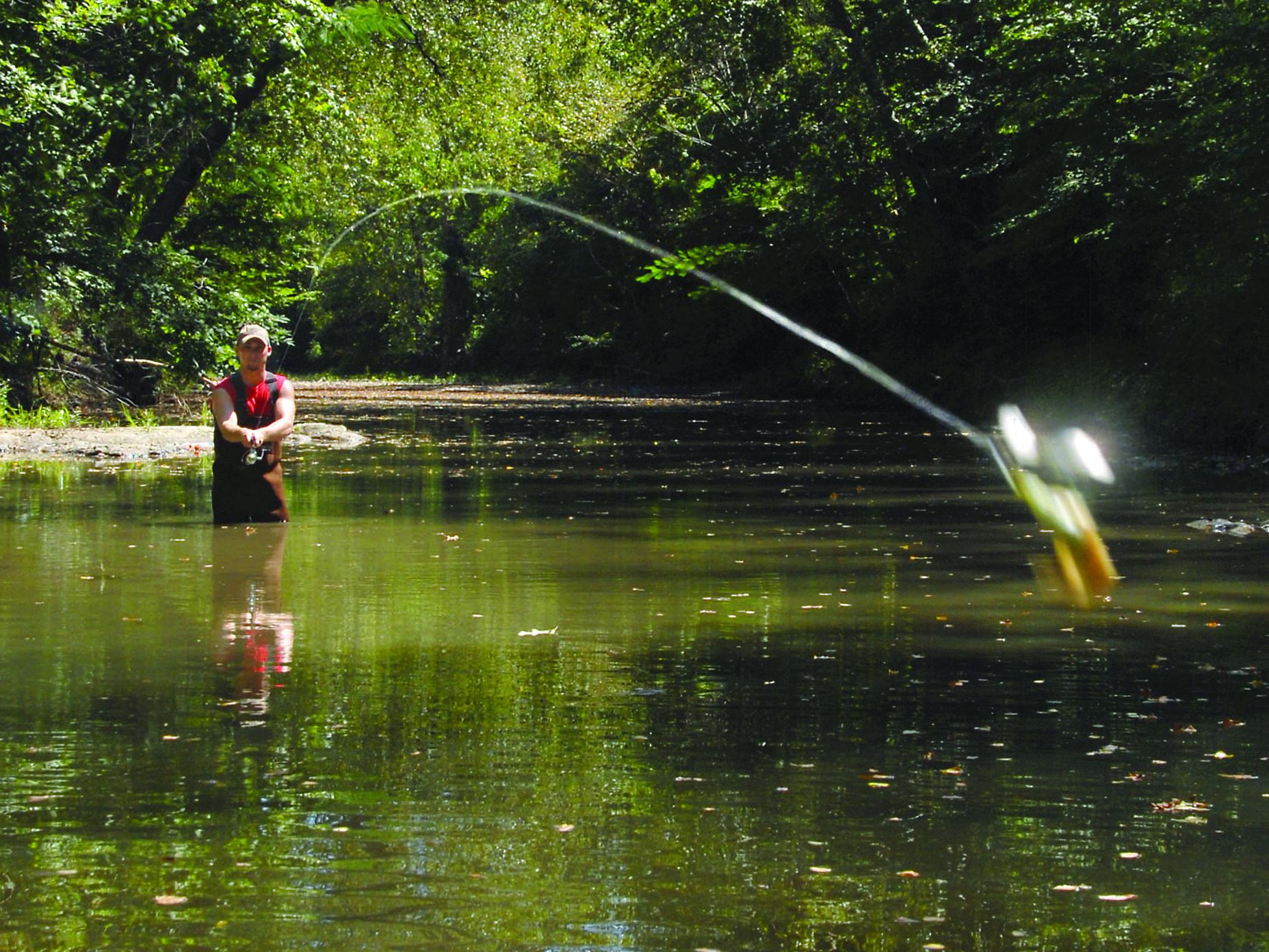 A fisherman in rubber waders stands in a small, quiet stream and casts a lure toward the viewer.