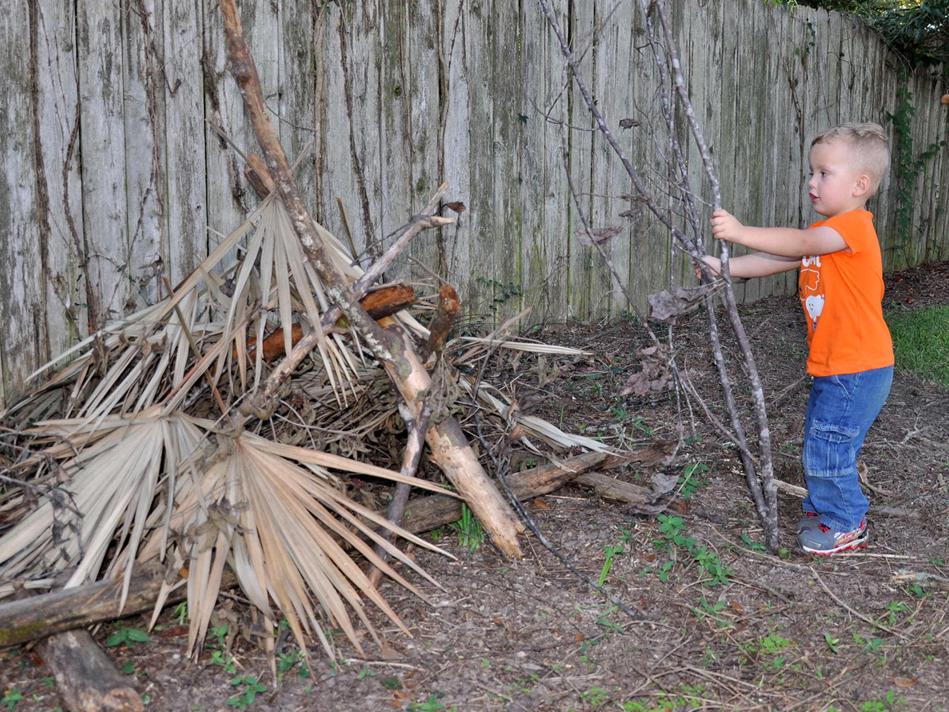 Preschool boy places a dead tree branch on a pile of limbs and leaves located beside an old, wooden privacy fence.