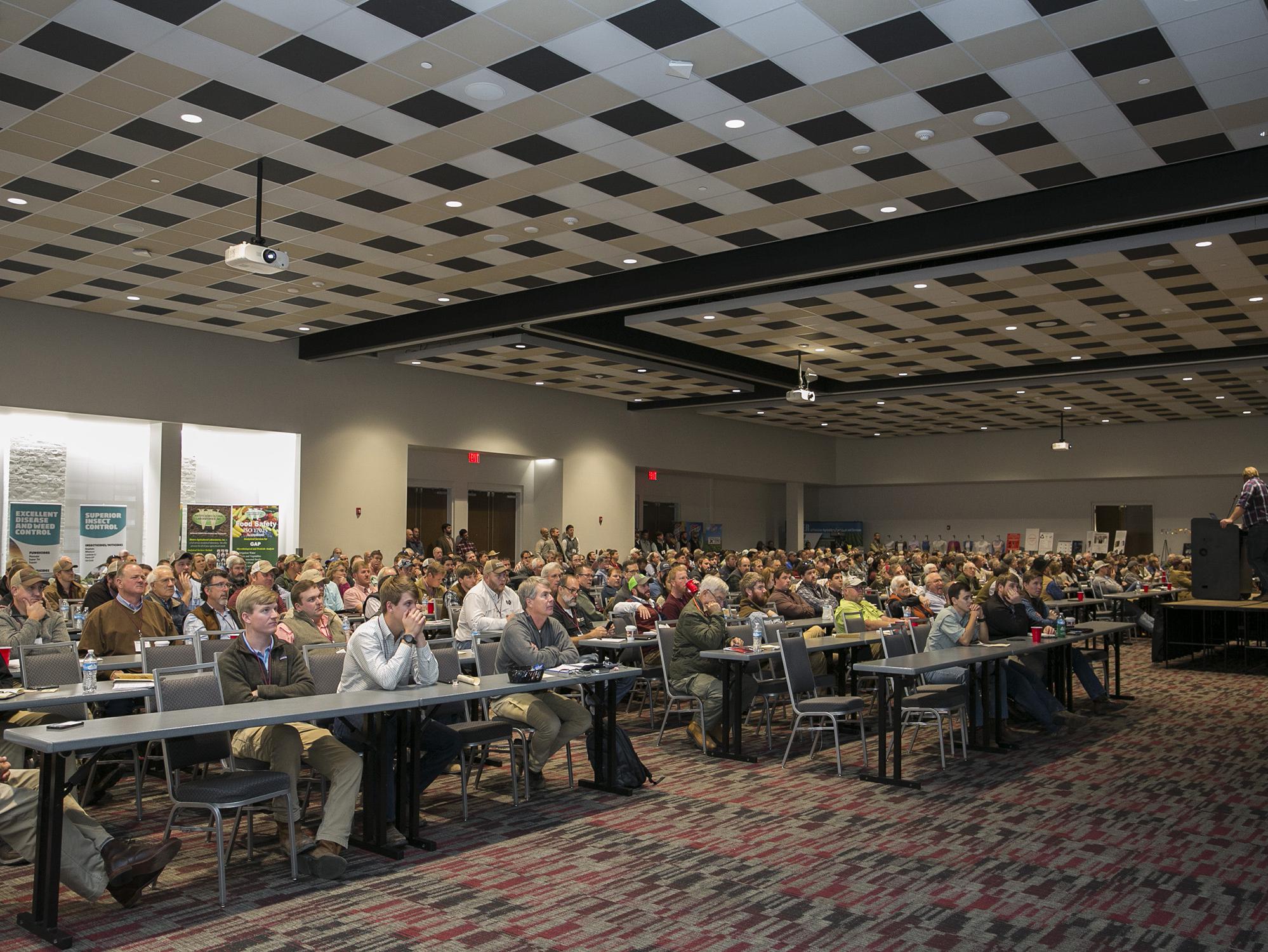 Hundreds of people in rows form an audience for a speaker in a large, open room.