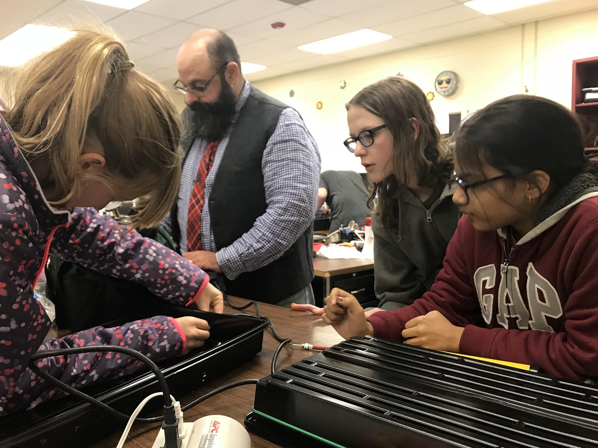 Two middle-school-aged girls and a middle-aged male teacher look on at another girl poking a hole in the side of a black tray for a science project.