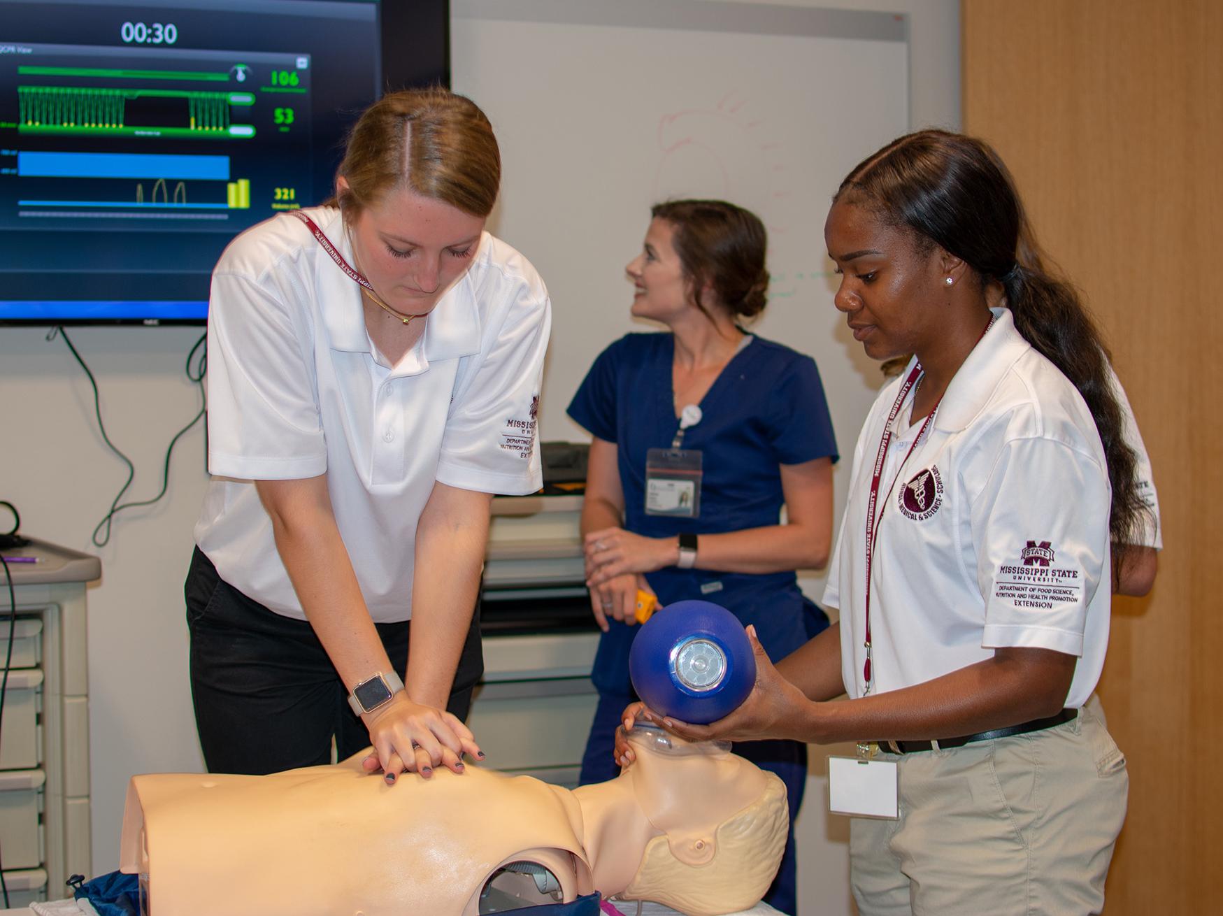 One teenager clasps both of her hands to press on a training manikin’s chest, while another female student holds a ventilator mask over its face. A nurse in the background watches a large wall-mounted monitor.