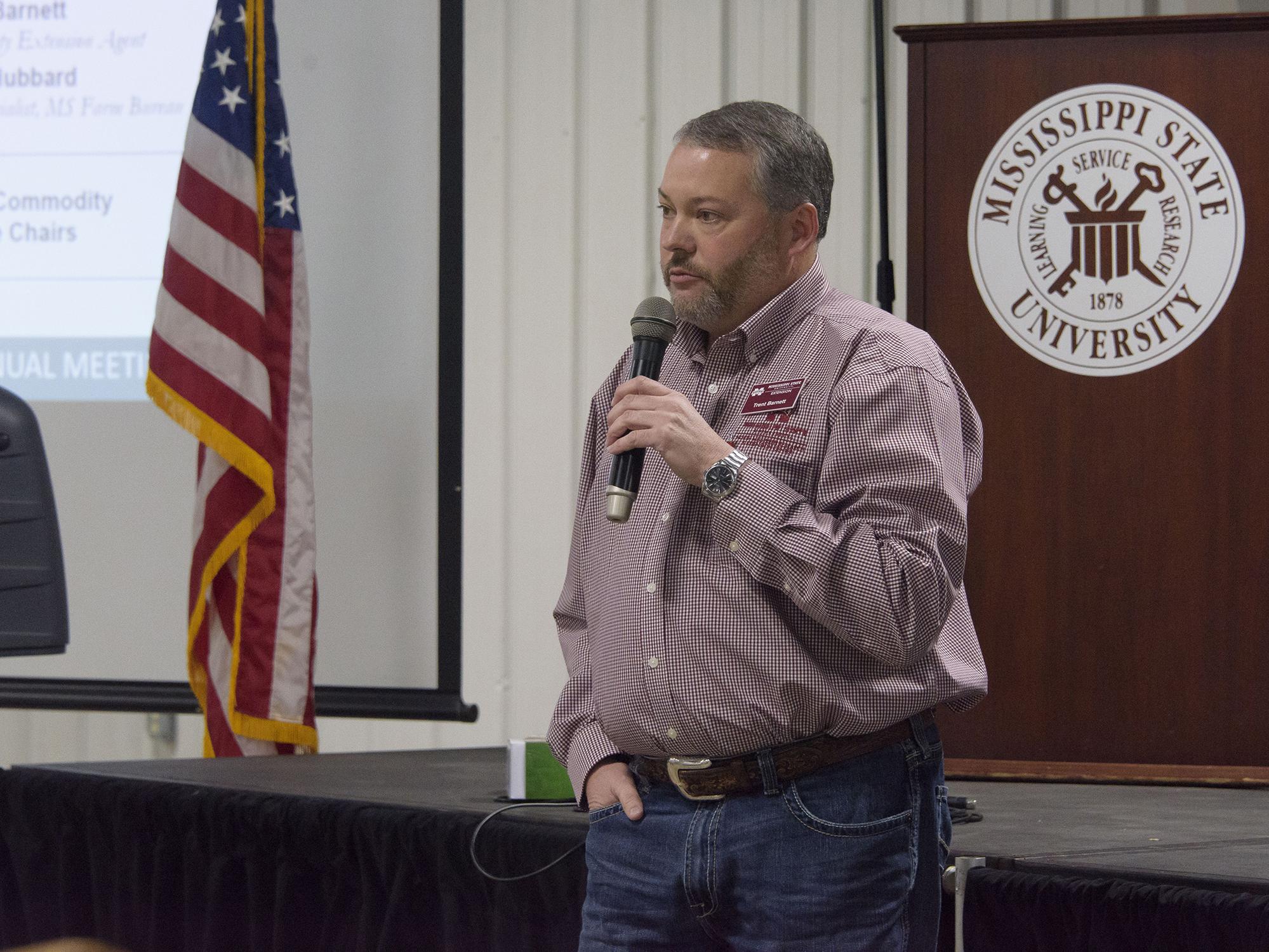 Man speaks into a handheld microphone. Background includes the Mississippi State University seal and an American flag.