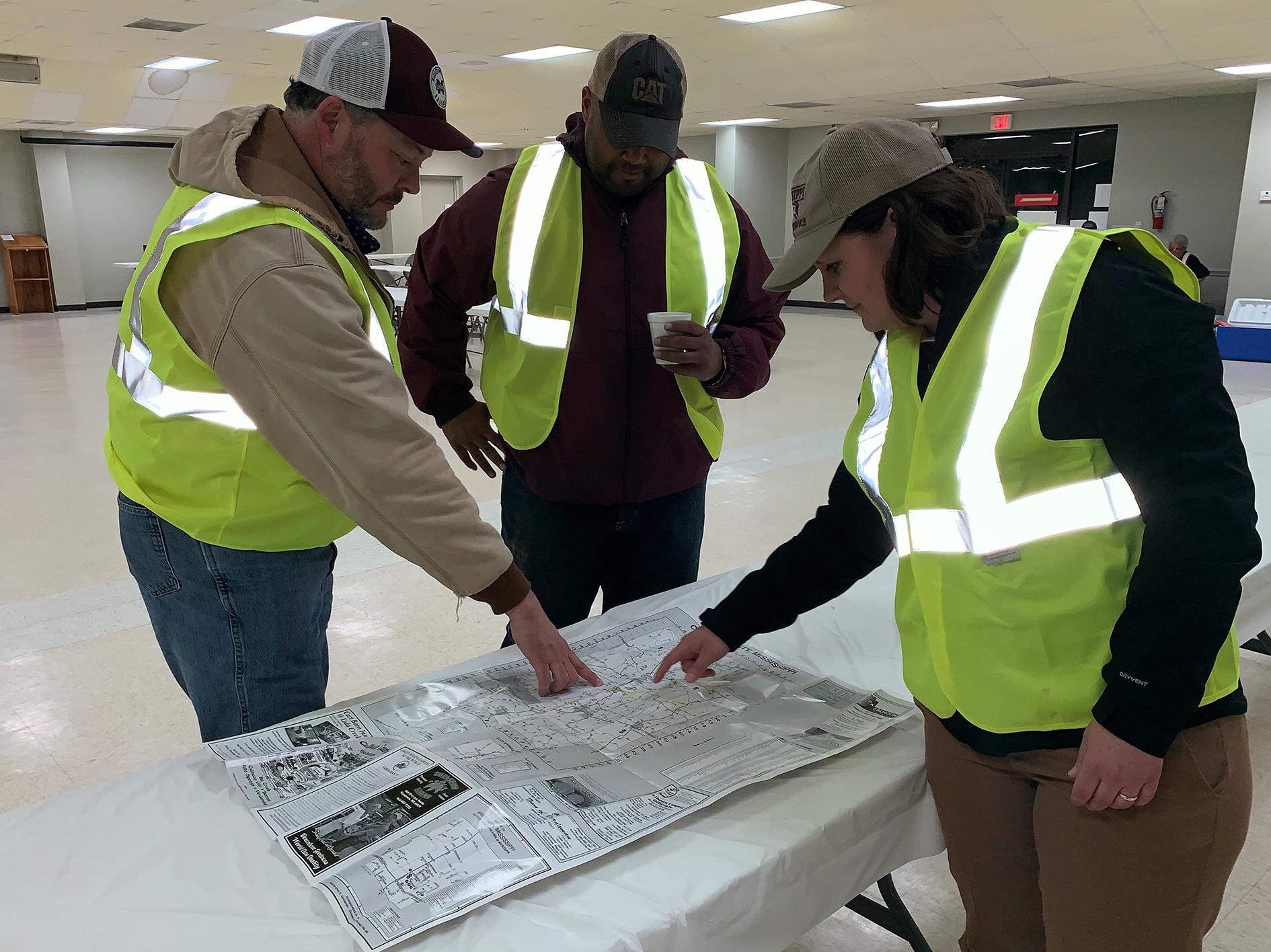 Two men and a woman, all wearing baseball caps and reflective yellow vests, look at a roadmap spread out on a table in a large, empty, well-lit room.