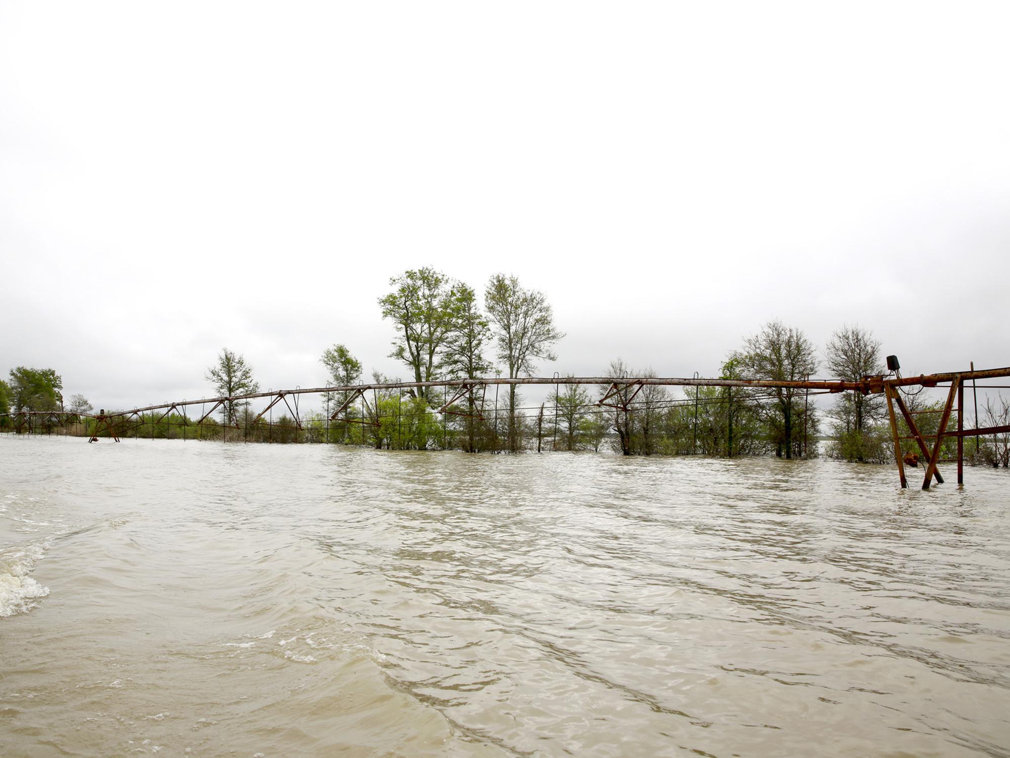 The rusty poles of an overhead, pivot irrigation system and a thin row of trees rise from the waves and gray floodwaters under a bleak sky.