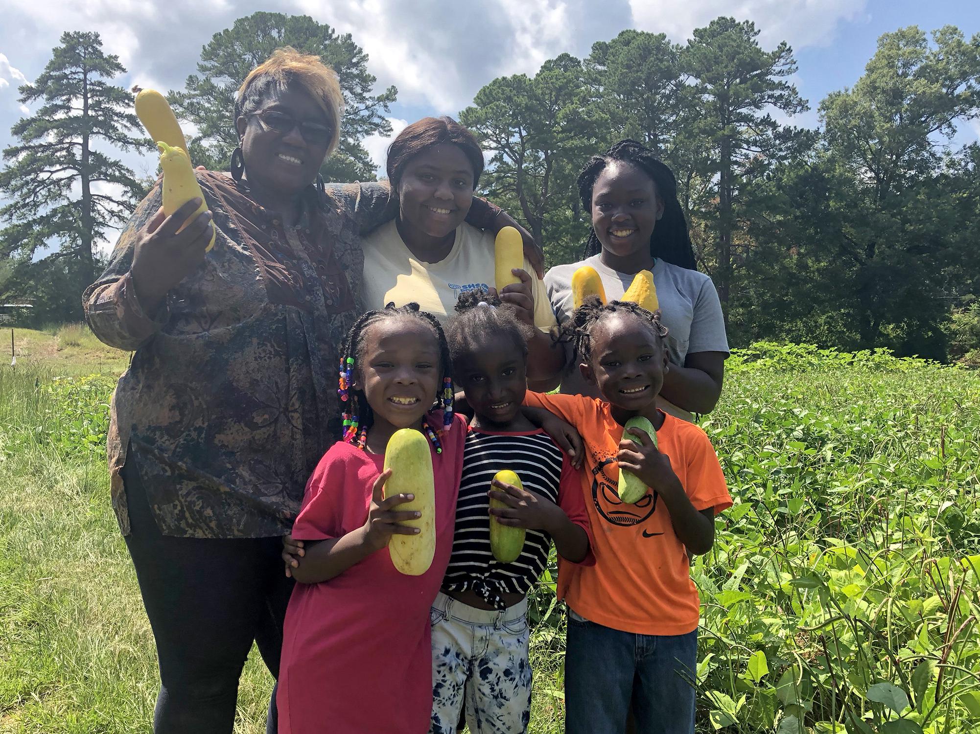 Two women and four children stand in a vegetable garden while holding yellow squash.