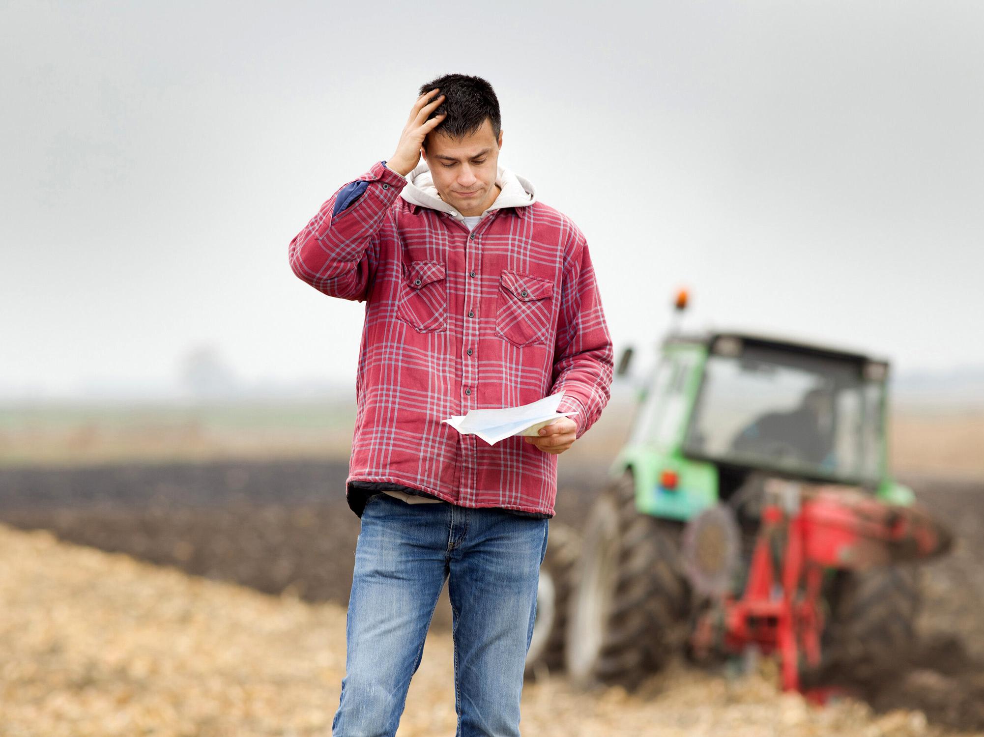 A farmer stands with a tractor in the background looking at a document and holding a hand to his head in worry.
