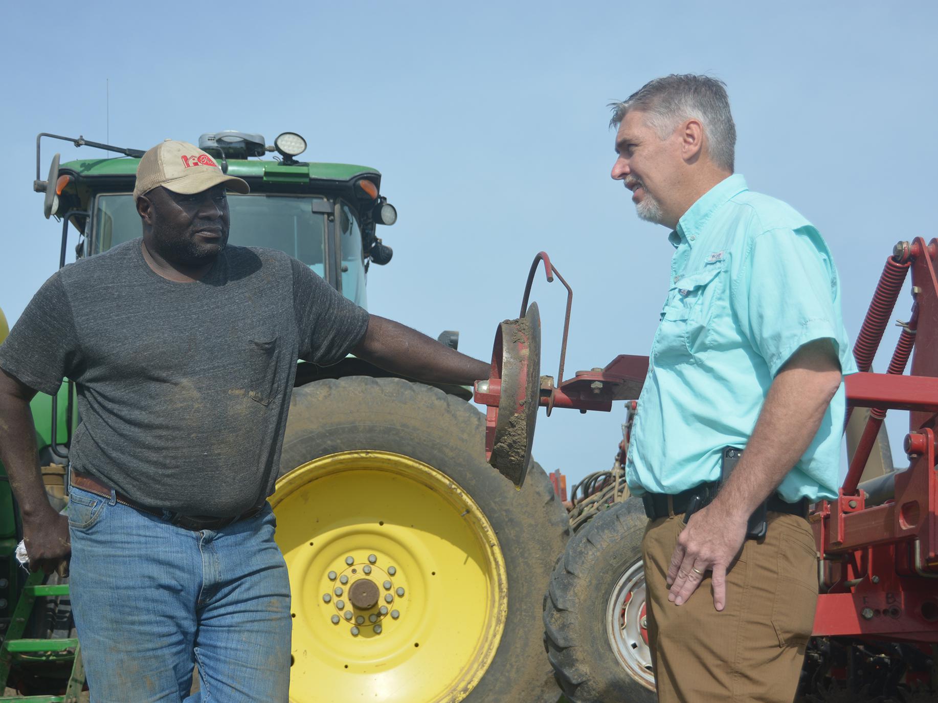 Two men facing each other in conversation and standing beside a tractor and equipment with a clear, blue sky overhead.