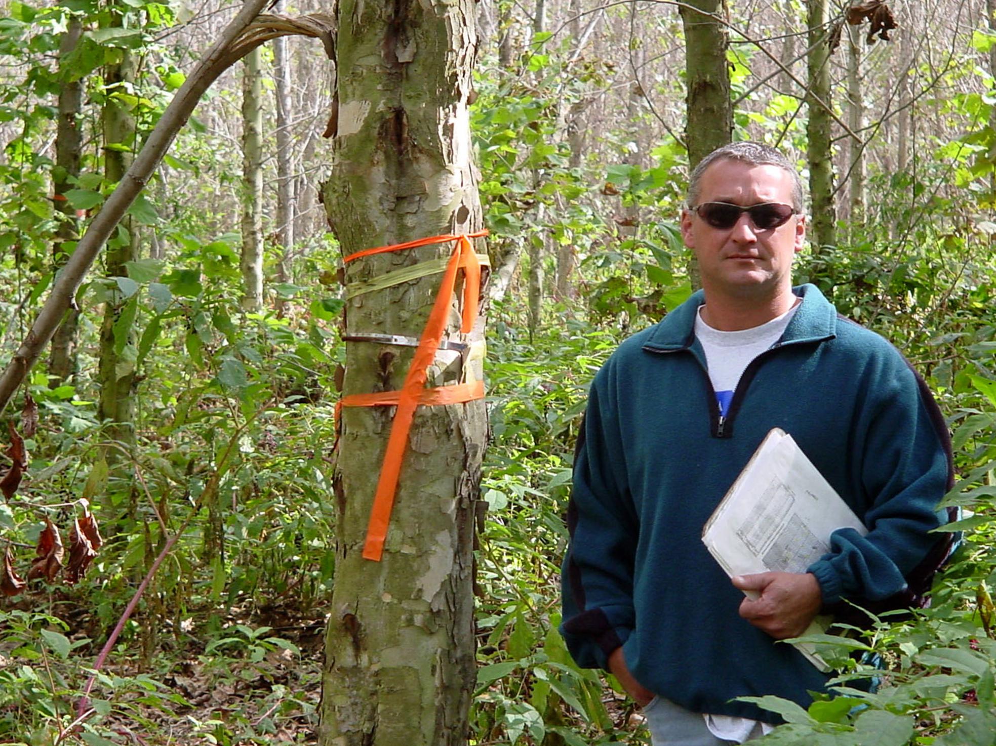 Man holding a clipboard looks at camera while standing in a wooded area beside a tree with orange ribbons tied around the trunk at shoulder height.
