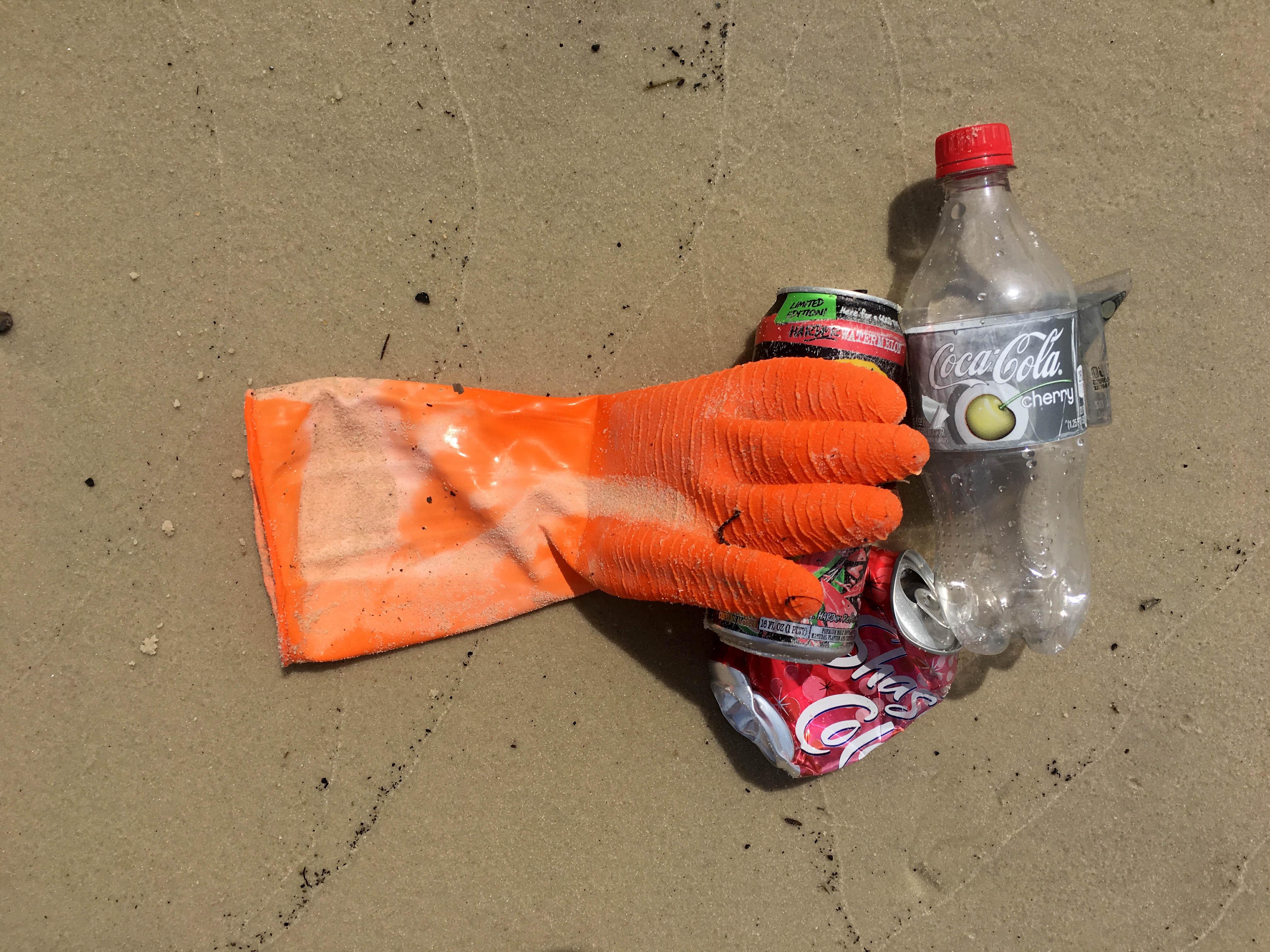 Small pile containing a rubber glove, an empty plastic bottle and two aluminum cans on wet sand.