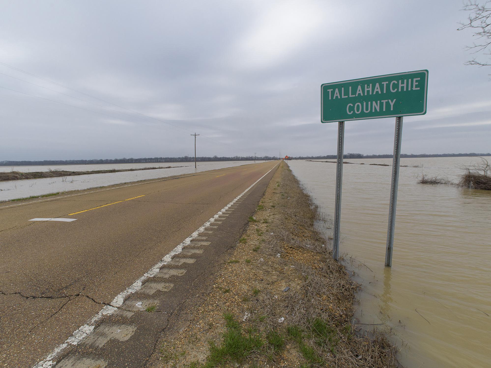 The view down a two-lane road with a wide expanse of water on each side and nearly touching the road. A road sign marks the Tallahatchie County line.