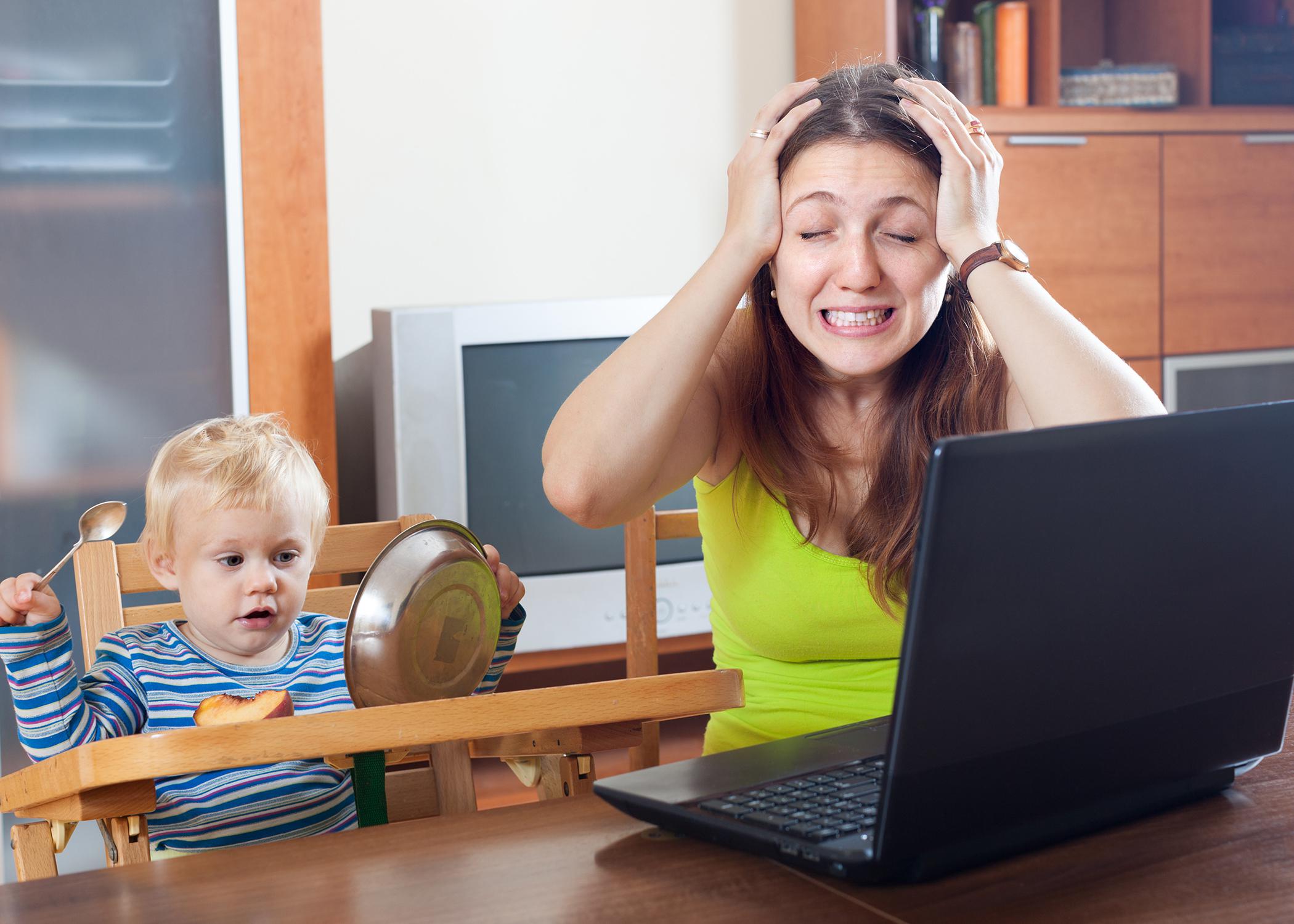 A young mother grabs her head in frustration while working from home on a laptop computer as her baby looks on. 