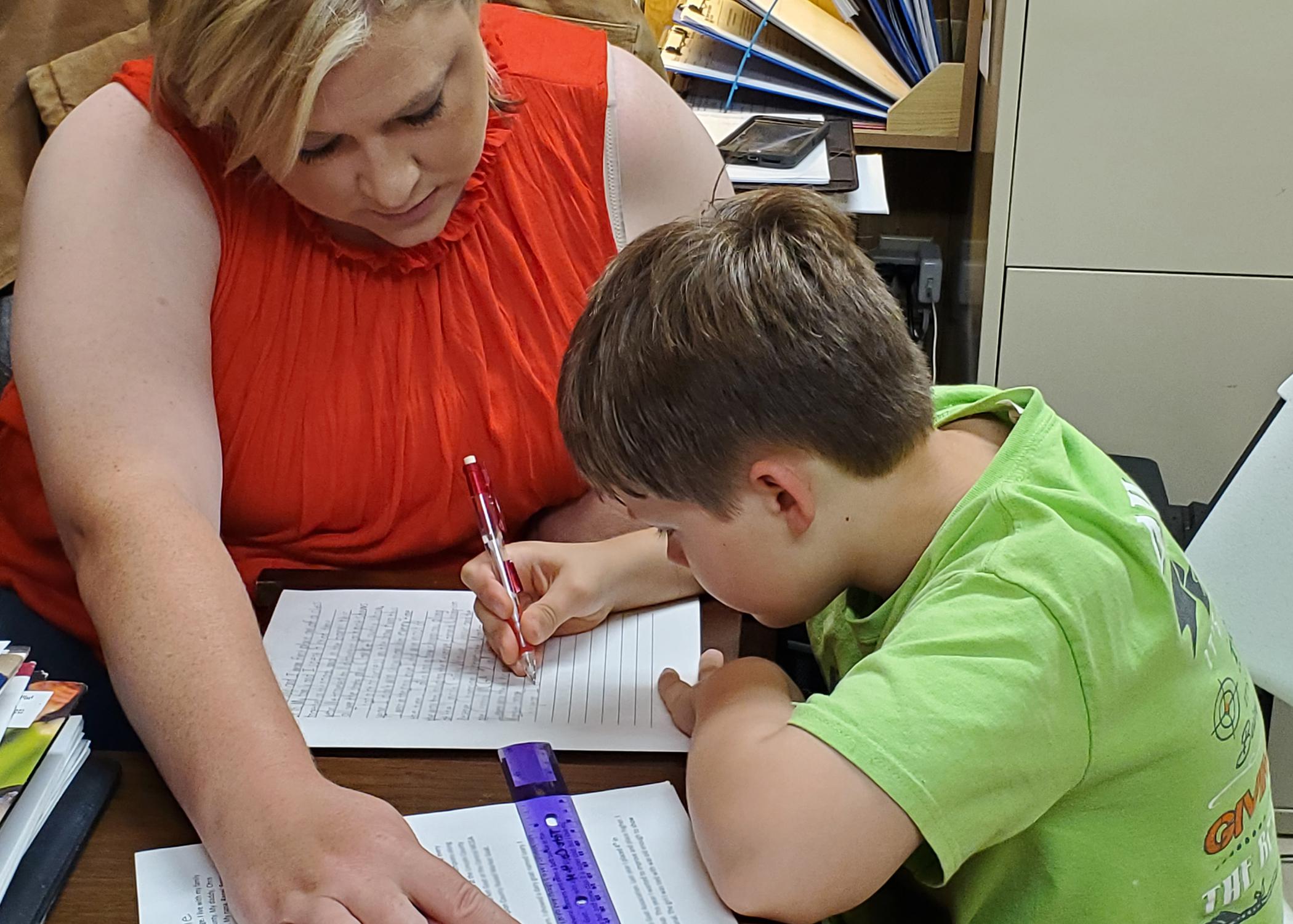 A woman looks on while a young boy writes on a piece of paper.