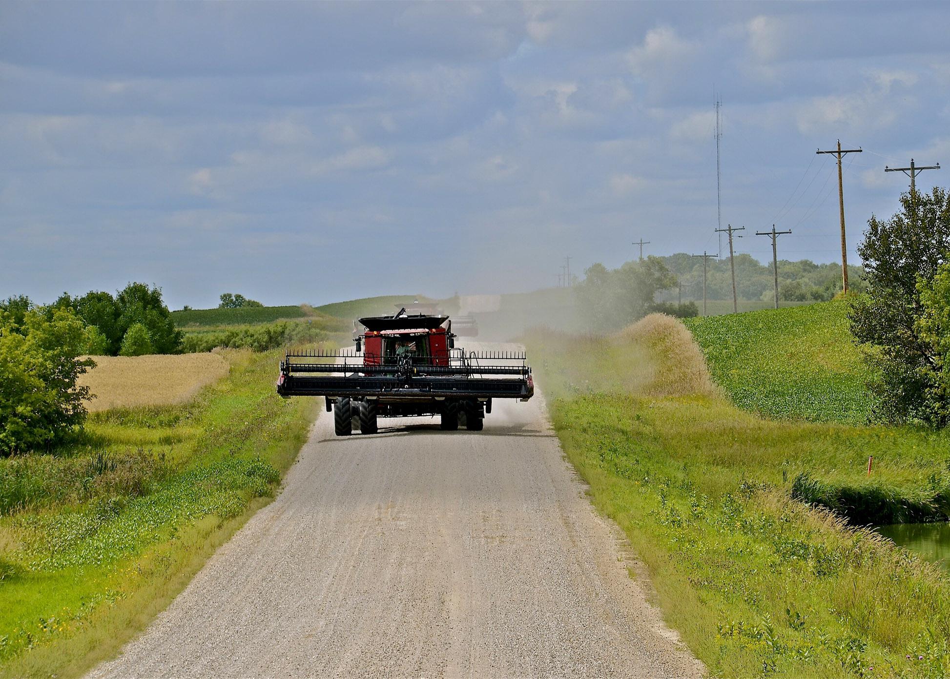 A red combine drives down a gravel road with farmland on both sides.