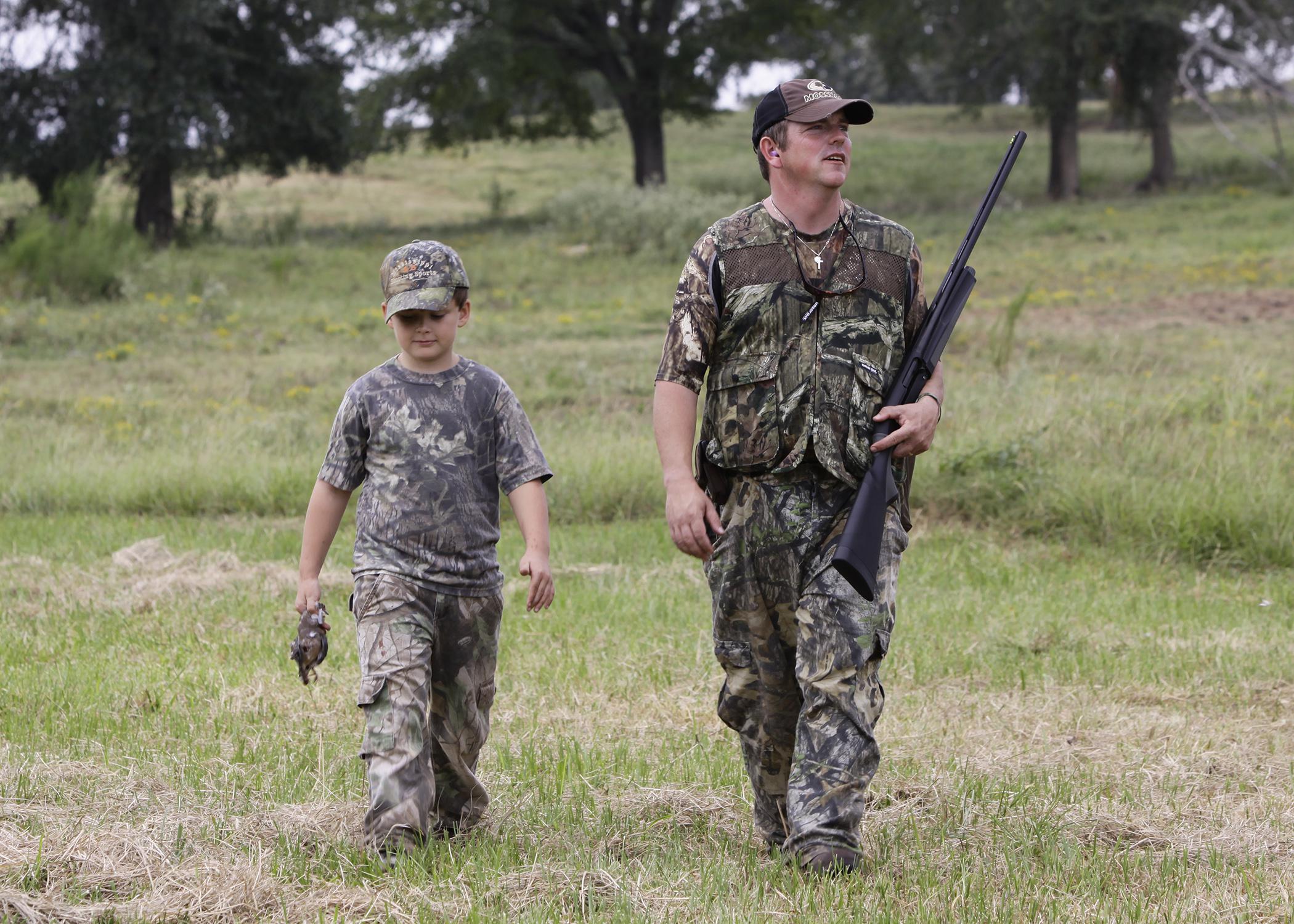 A man holding a shotgun and a boy dressed in camouflage walk in a grassy meadow.