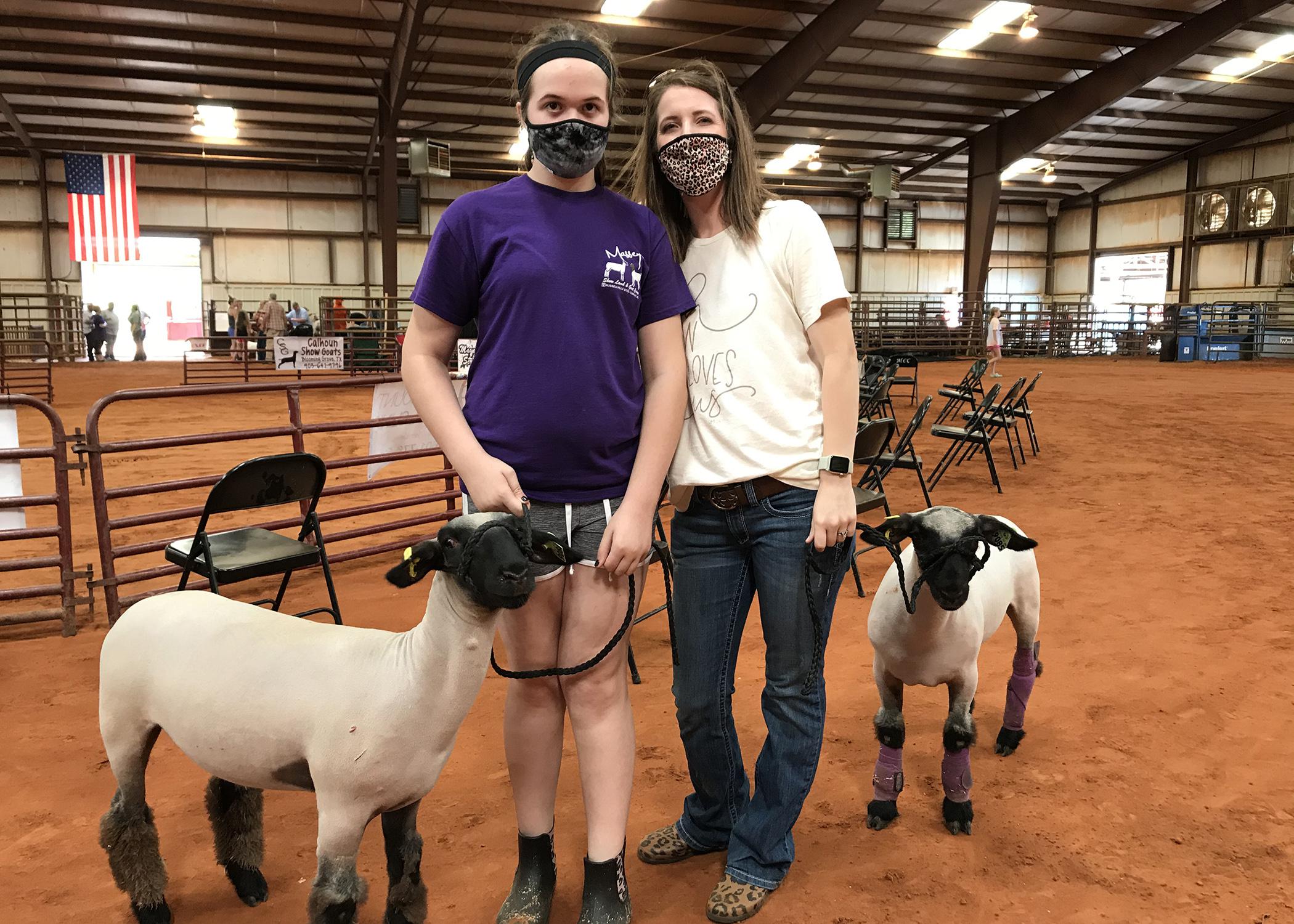 A daughter and her mother wearing masks in a rodeo building with two lambs leashed beside them.