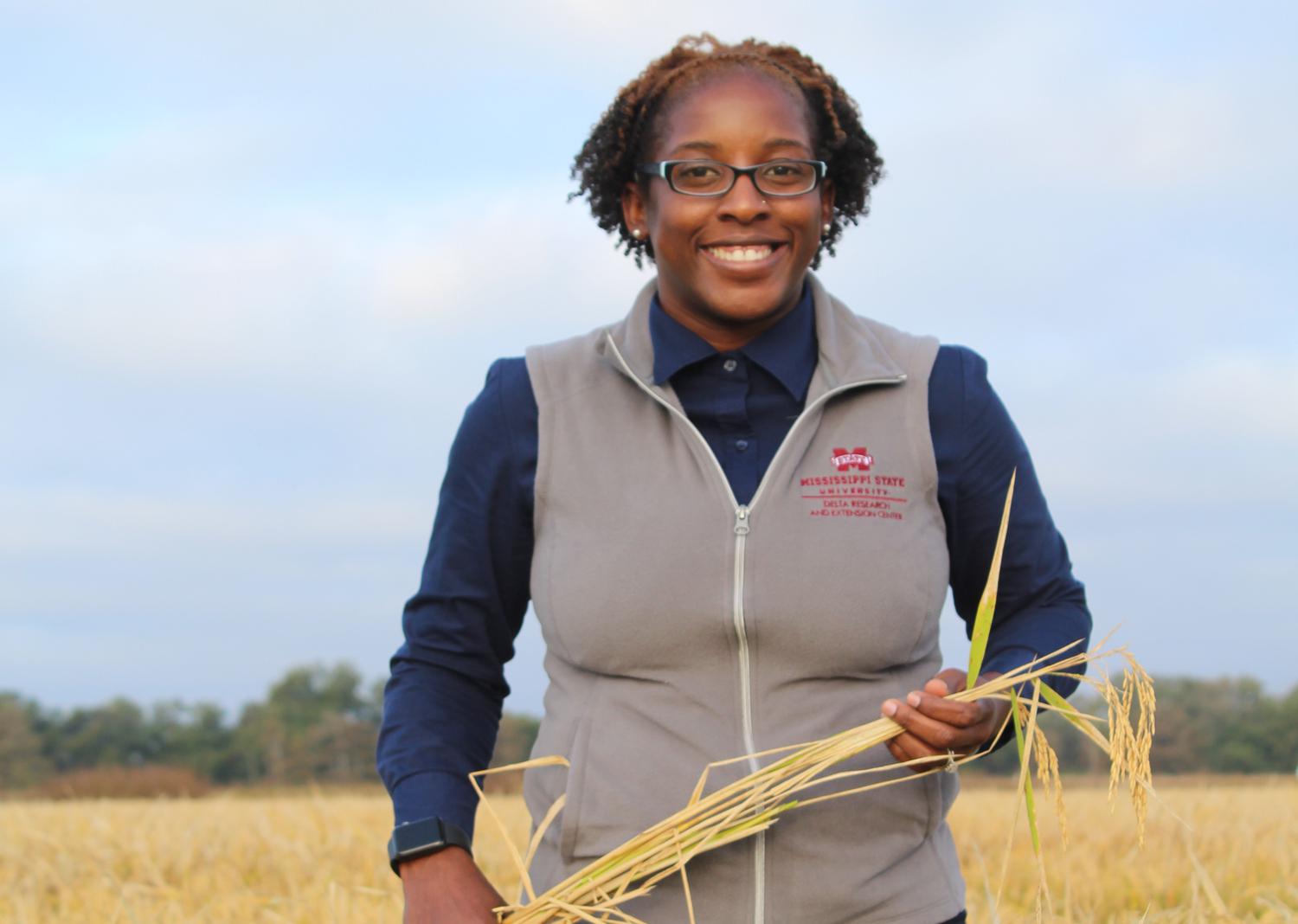A woman holds a stalk of grain while standing in a field.