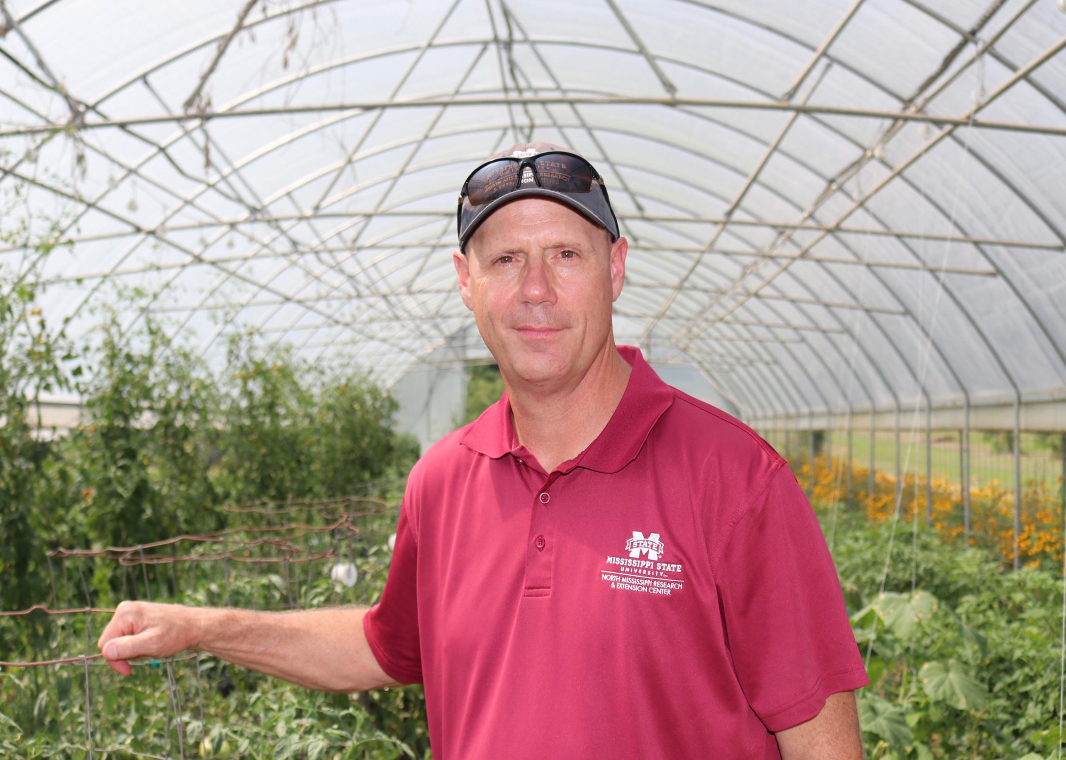 Man in a maroon shirt and baseball cap in a greenhouse.