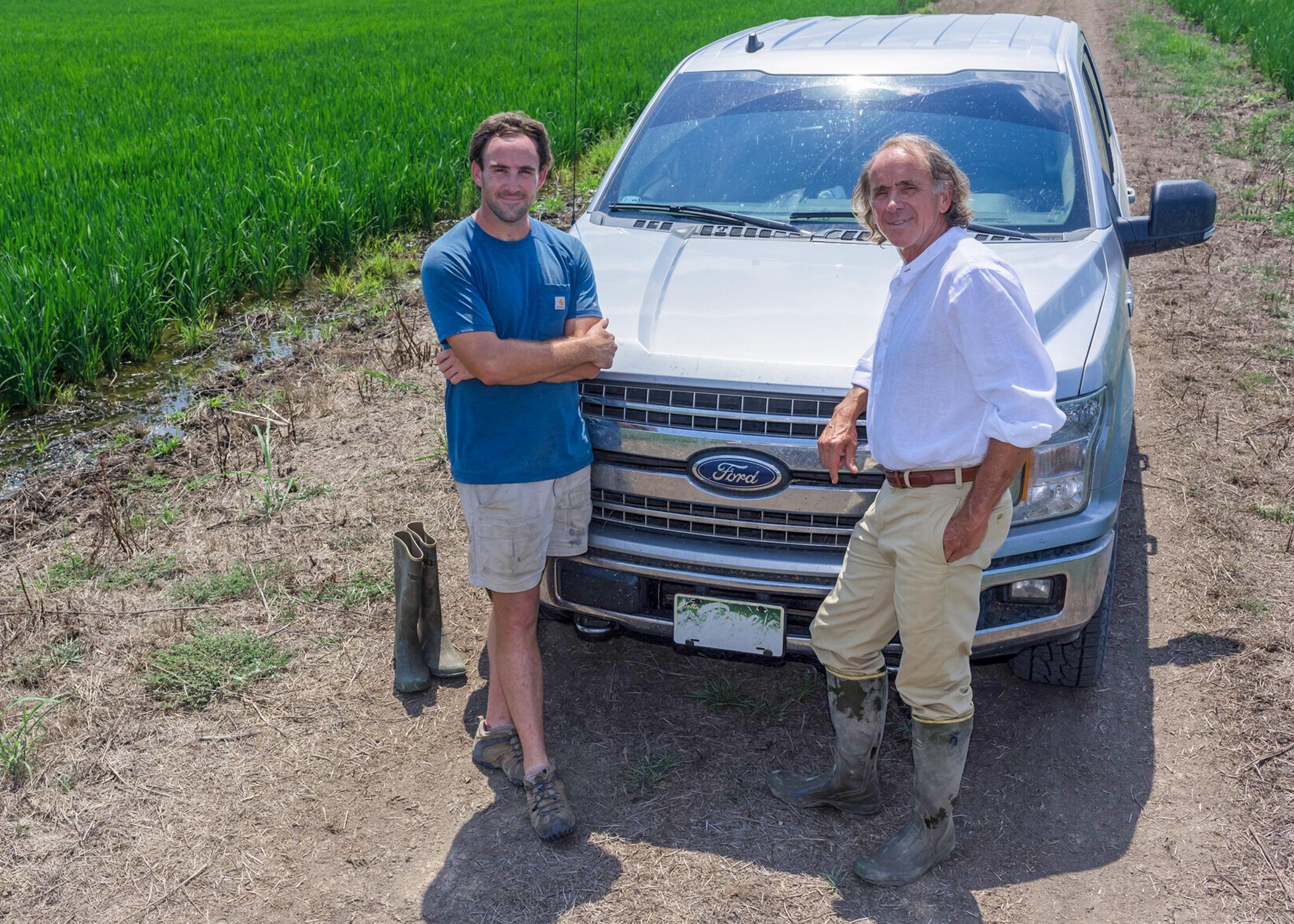Two men standing in front of a pickup truck parked in a rice field.