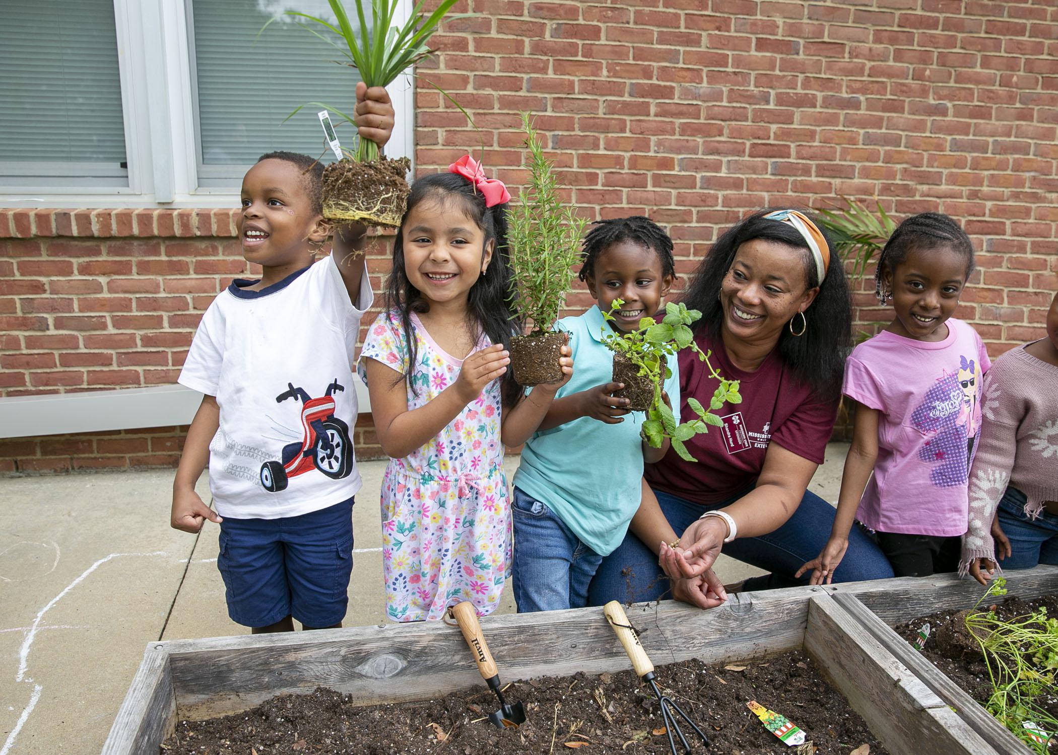 Four students stand in a row with their teacher and hold up the herbs they were about to plant in the raised bed.