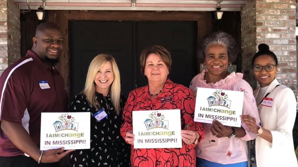 Four coalition members stand with Extension agent Alexis Hamilton. They hold small signs that read “I aim for change in Mississippi” with the AIM for CHangE logo.