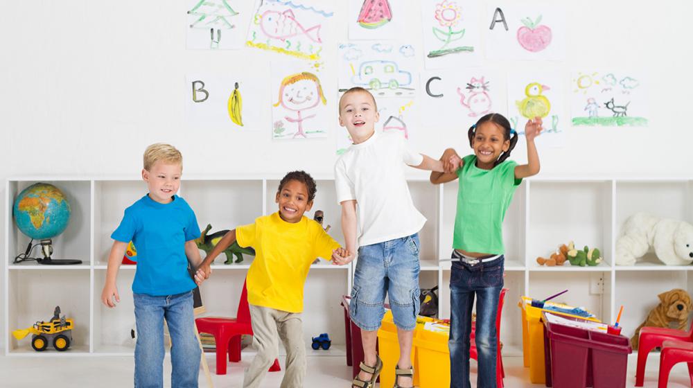 4 young children standing in a classroom, holding hands and jumping.