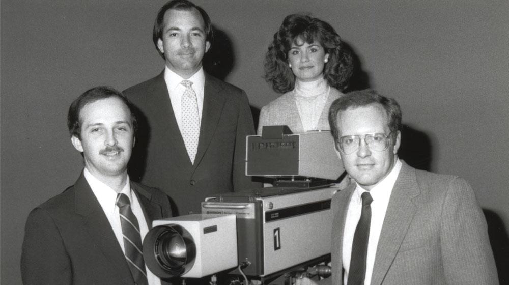 News crew, 3 men and a woman, posed around an old television camera.