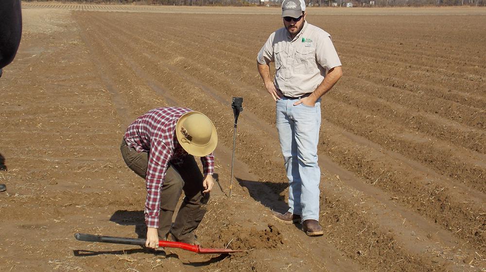 Woman using shovel in field to get soil sample while man looks on.
